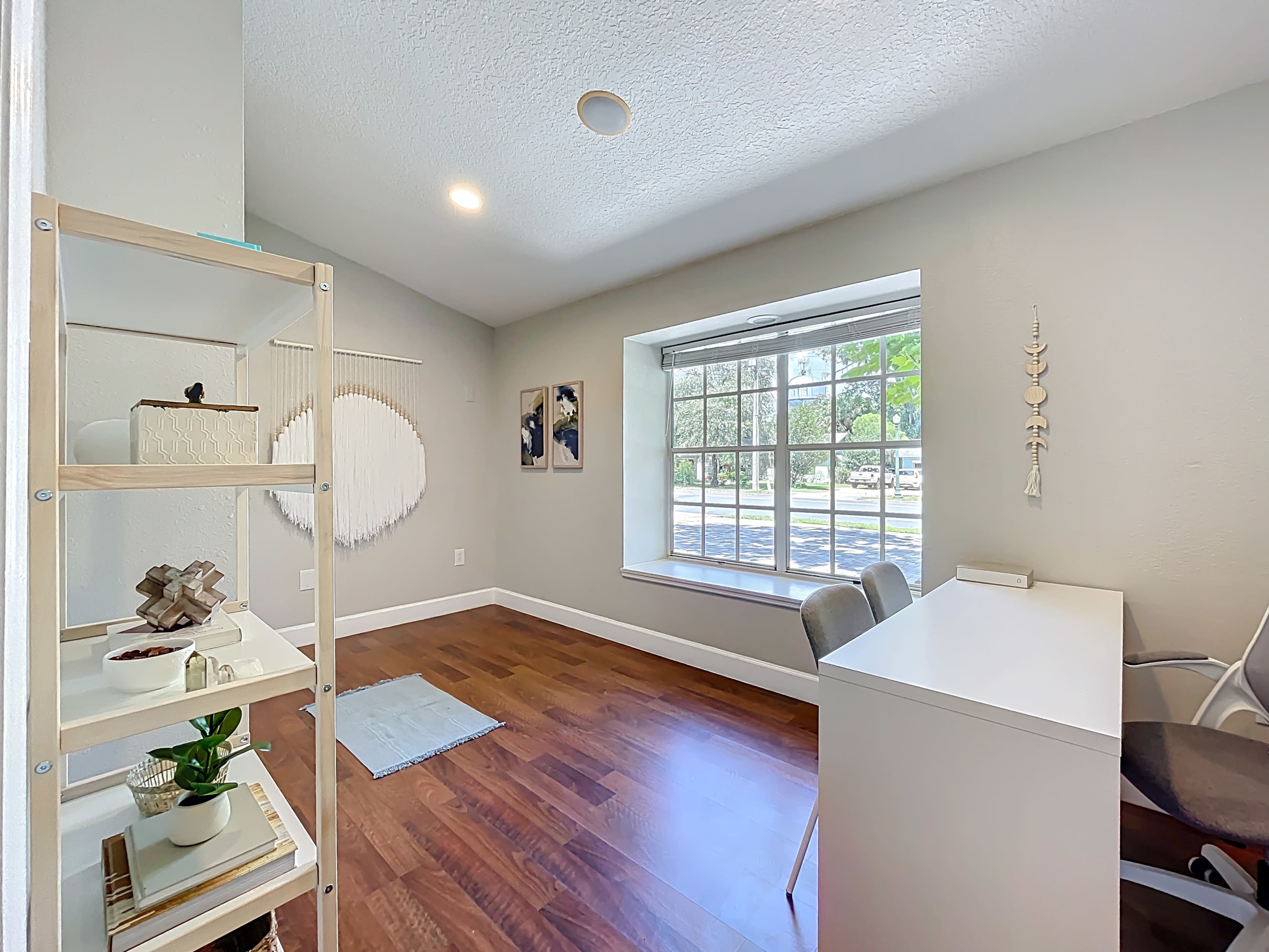 The image shows a bright, minimalist room featuring a desk, two chairs, and a window with a view of greenery outside.