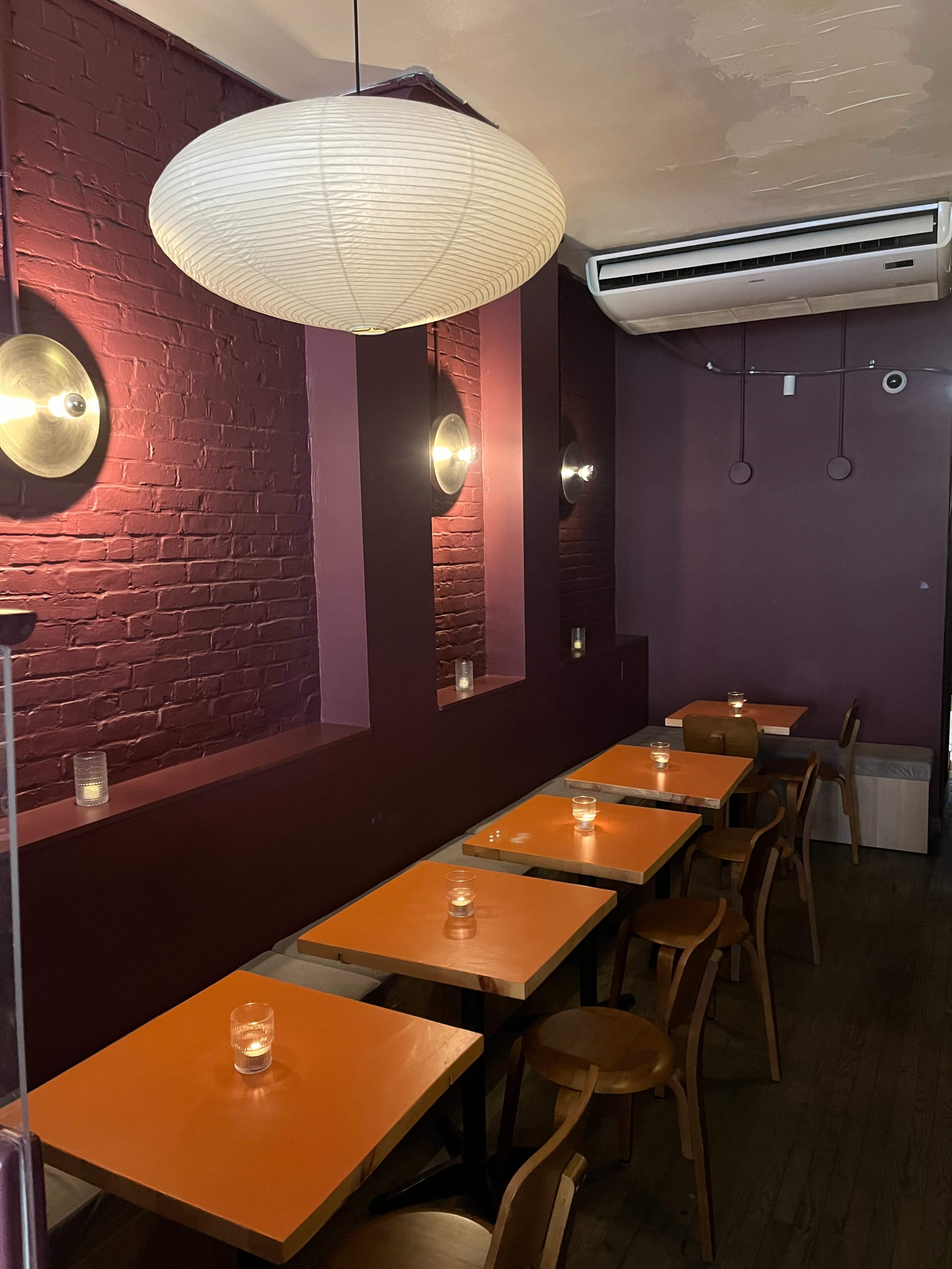The image shows a cozy café with purple walls, illuminated by wall-mounted lights and a large pendant lamp, featuring several wooden tables with orange tops and candles on them.