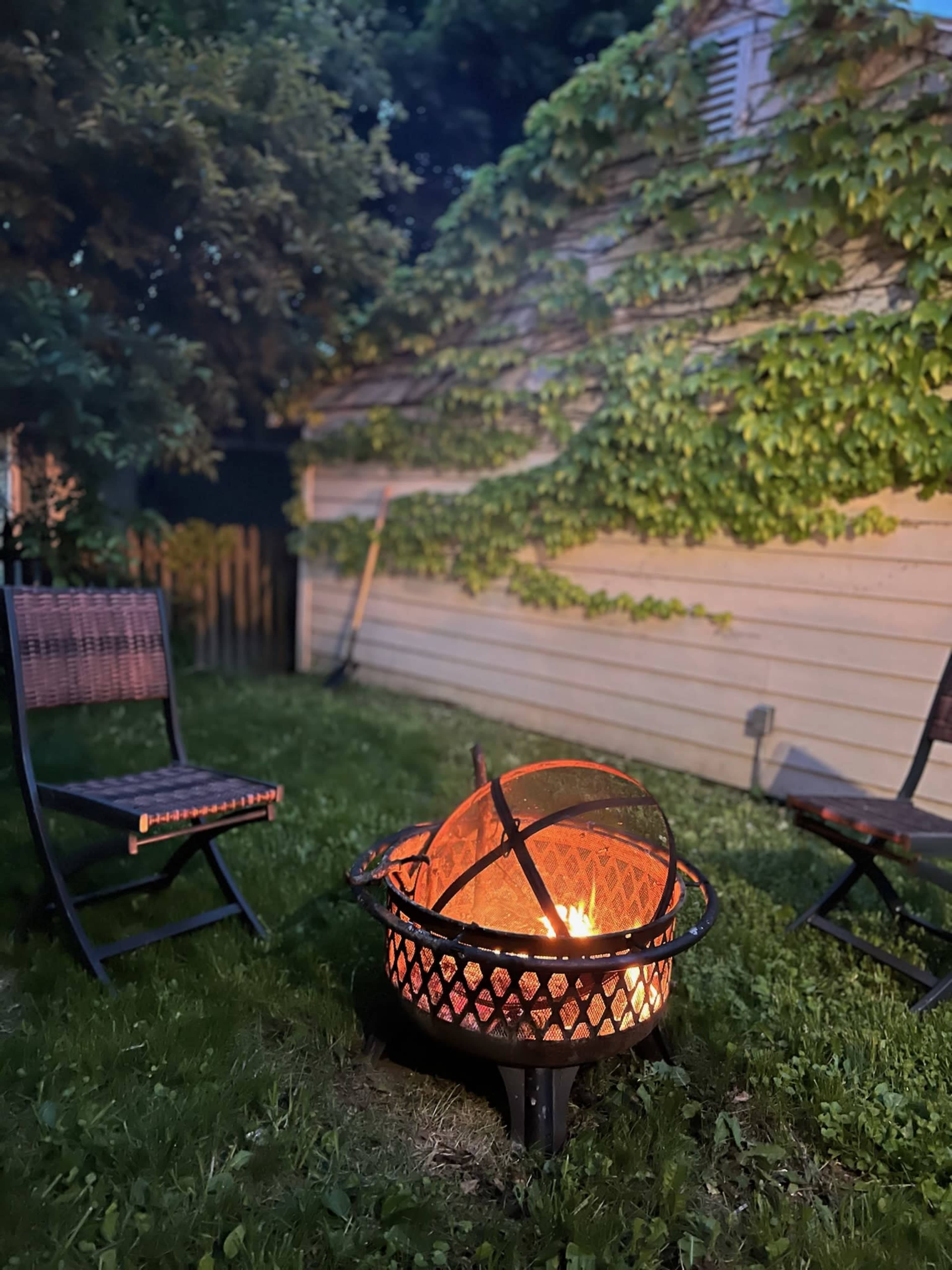 A fire pit with flames sits on grass between two wooden chairs in a garden surrounded by ivy-covered walls.