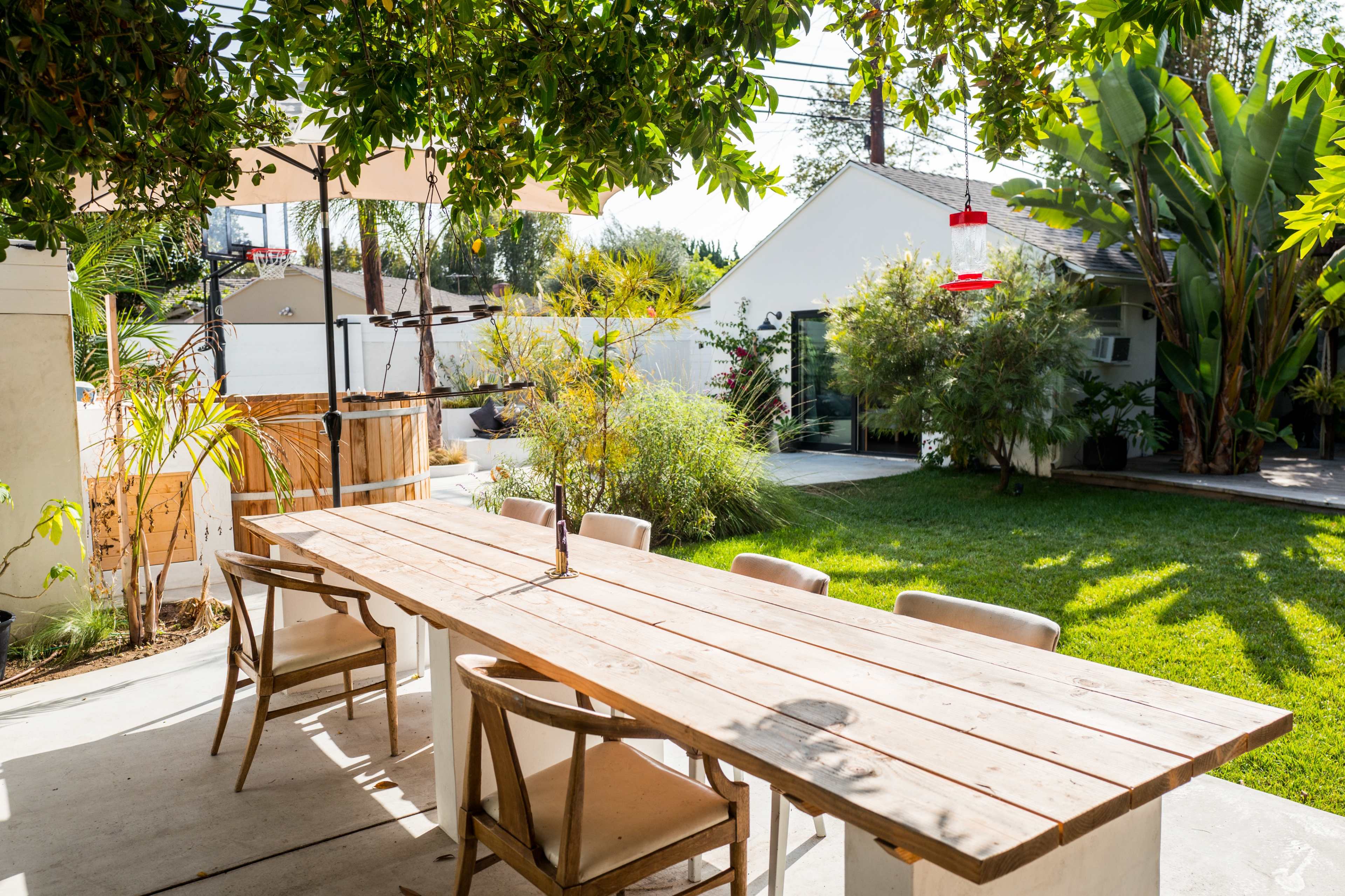 A spacious outdoor dining area with a long wooden table surrounded by chairs, set in a landscaped yard featuring greenery and a small building in the background.