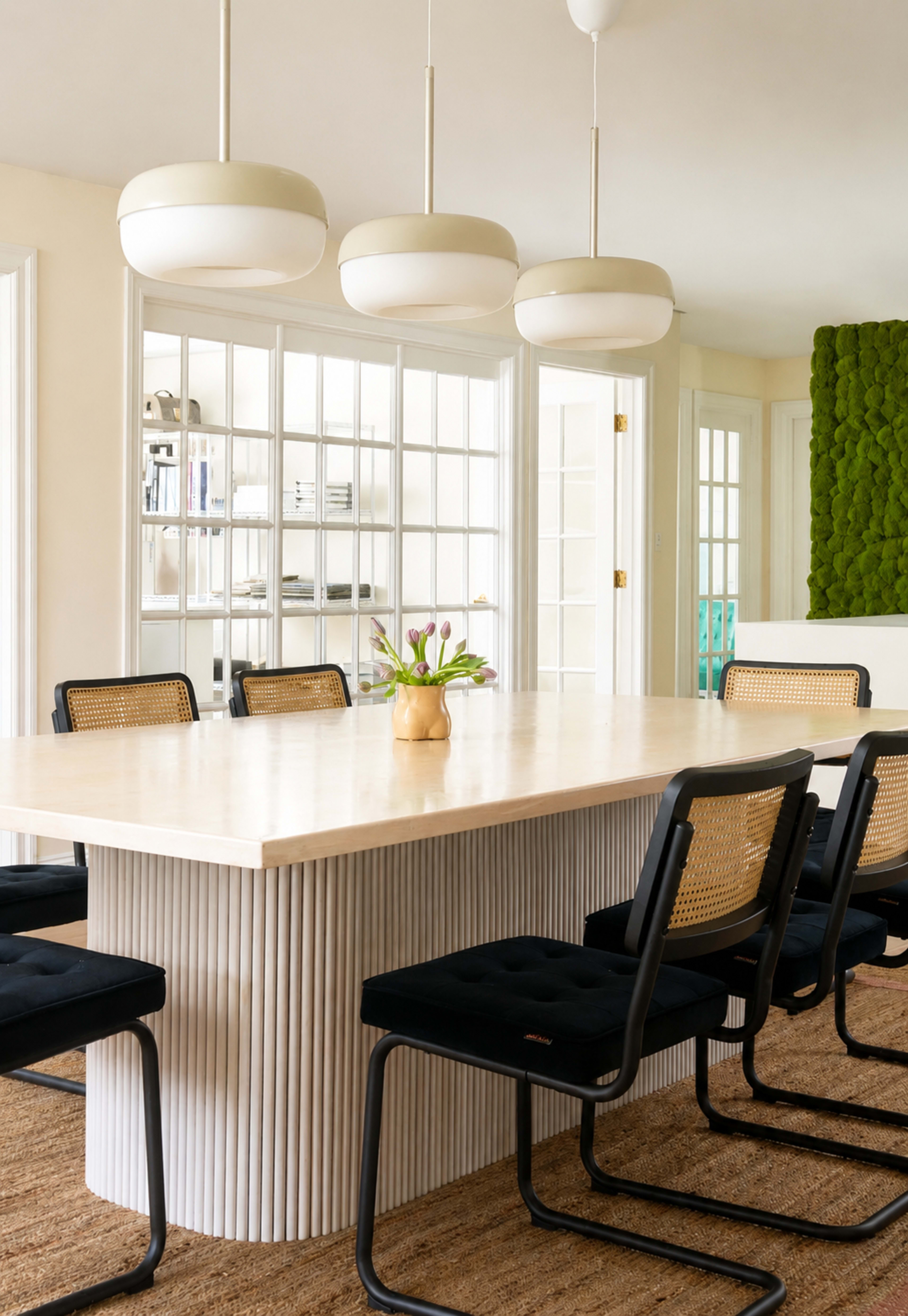A light-colored dining table with a textured base is surrounded by black chairs and illuminated by three pendant lights, set in a bright room with a green moss wall.