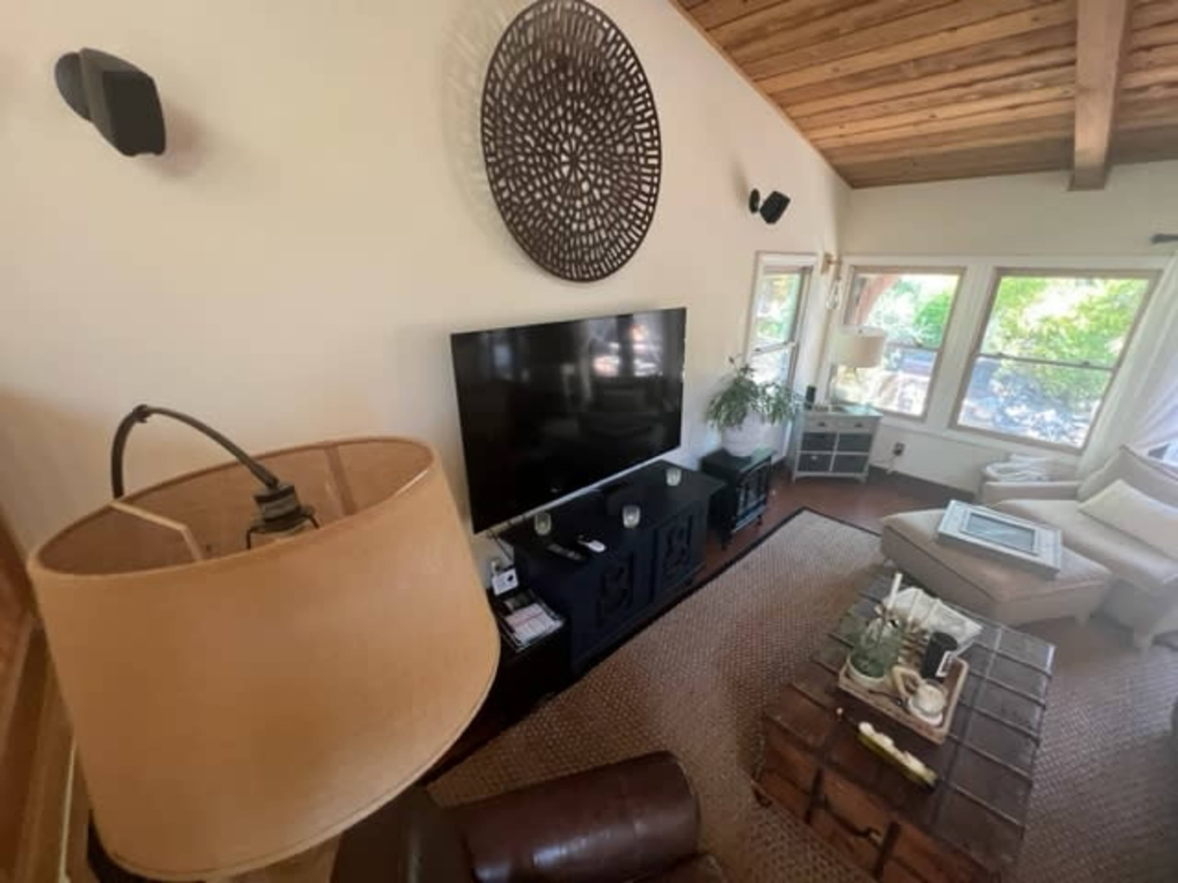 A cozy living room with a large television mounted on a wall, a wooden coffee table in front, and large windows allowing natural light to enter.