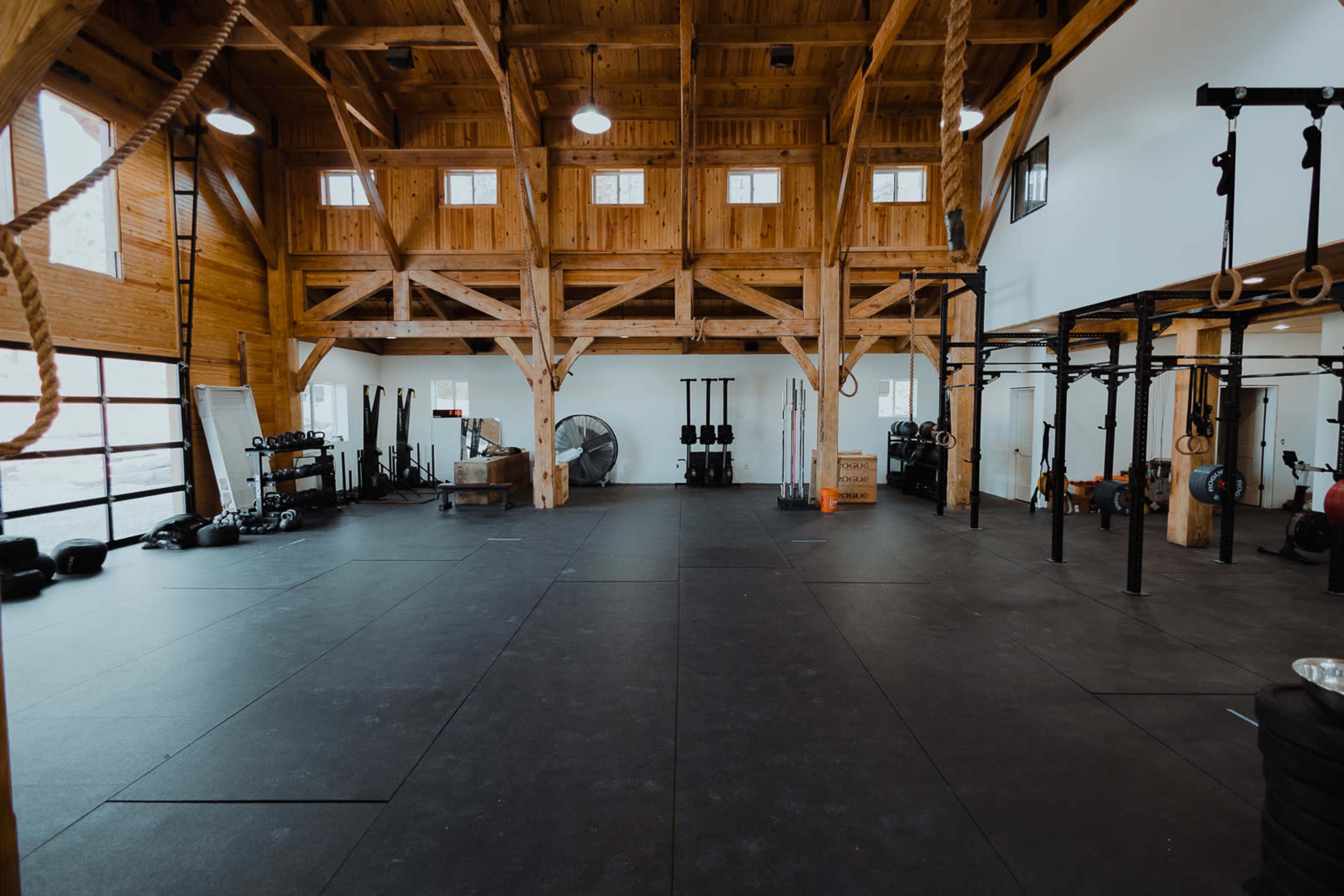 The image shows a spacious wooden gym interior with various exercise equipment, including weightlifting bars, ropes, and gymnastic rings.