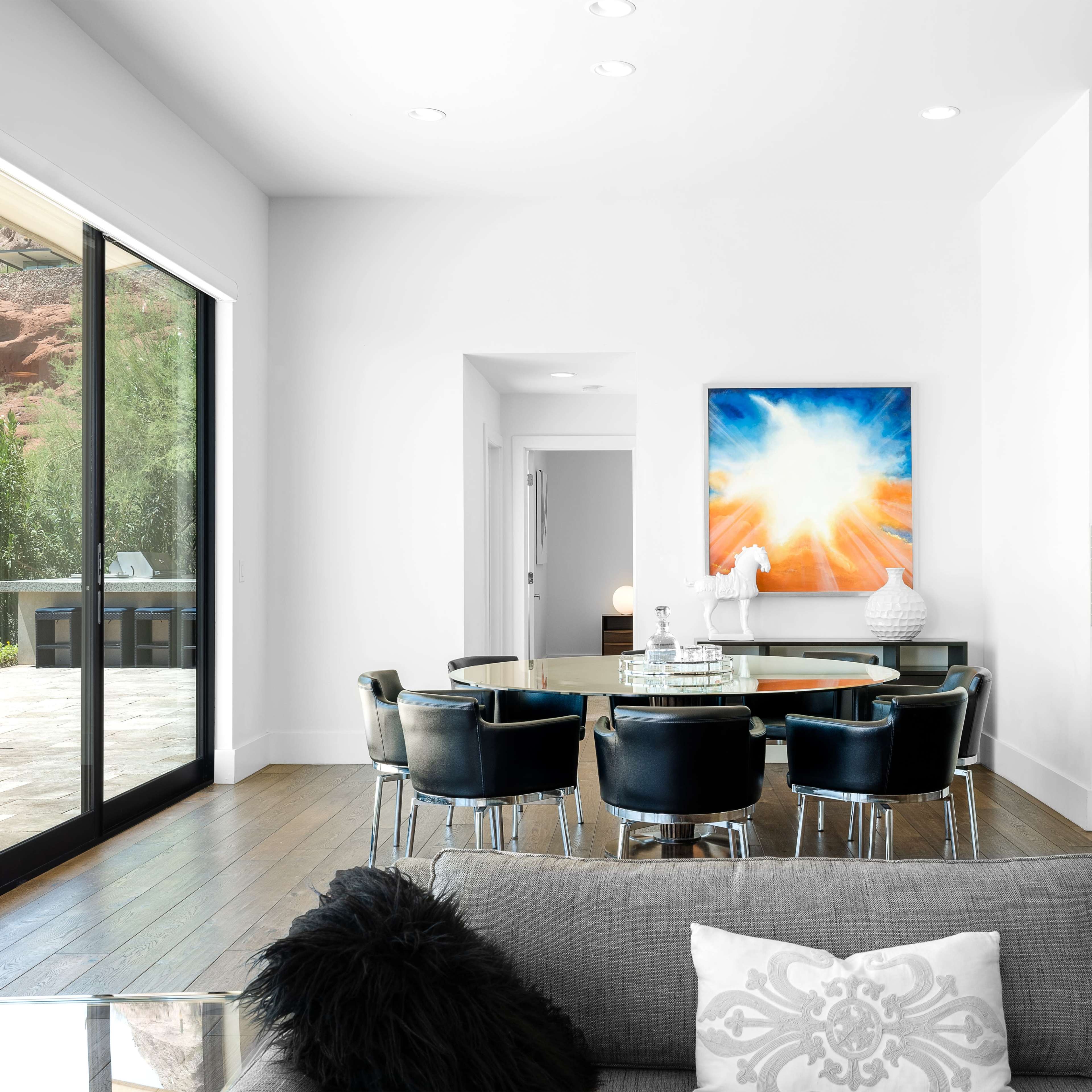 A modern dining area features a glass table surrounded by black chairs, with large sliding doors leading to an outdoor space and a vibrant artwork hanging on the wall.