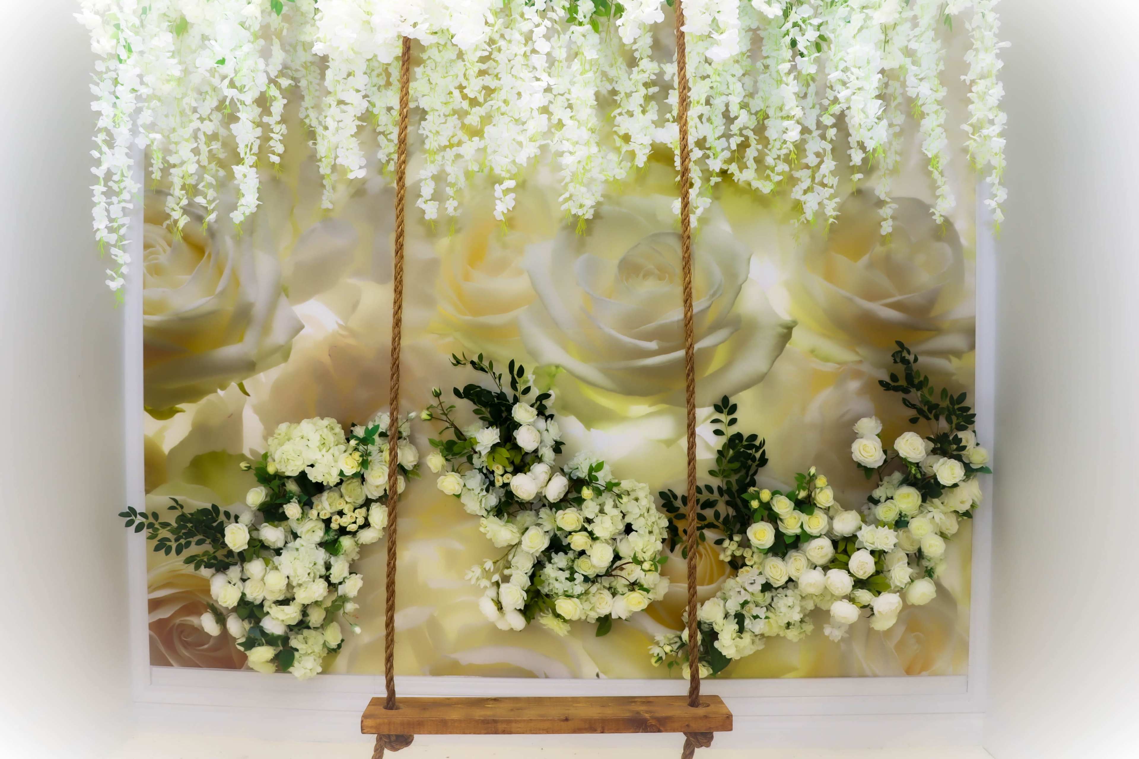 The image features a swing hanging from a floral arrangement with a backdrop of large white roses.