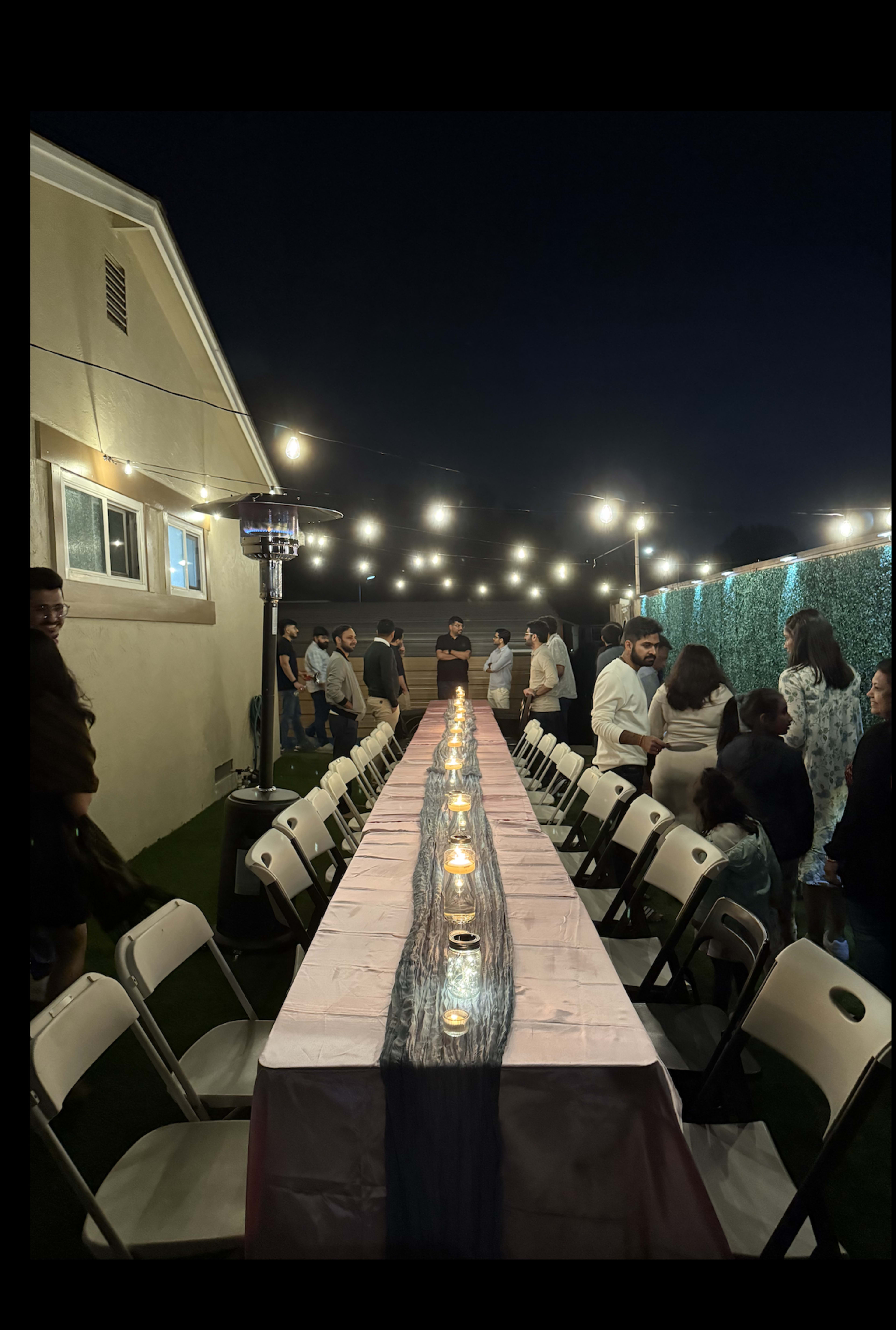 A long, illuminated table is set up outdoors for a gathering, surrounded by guests and soft overhead lights.