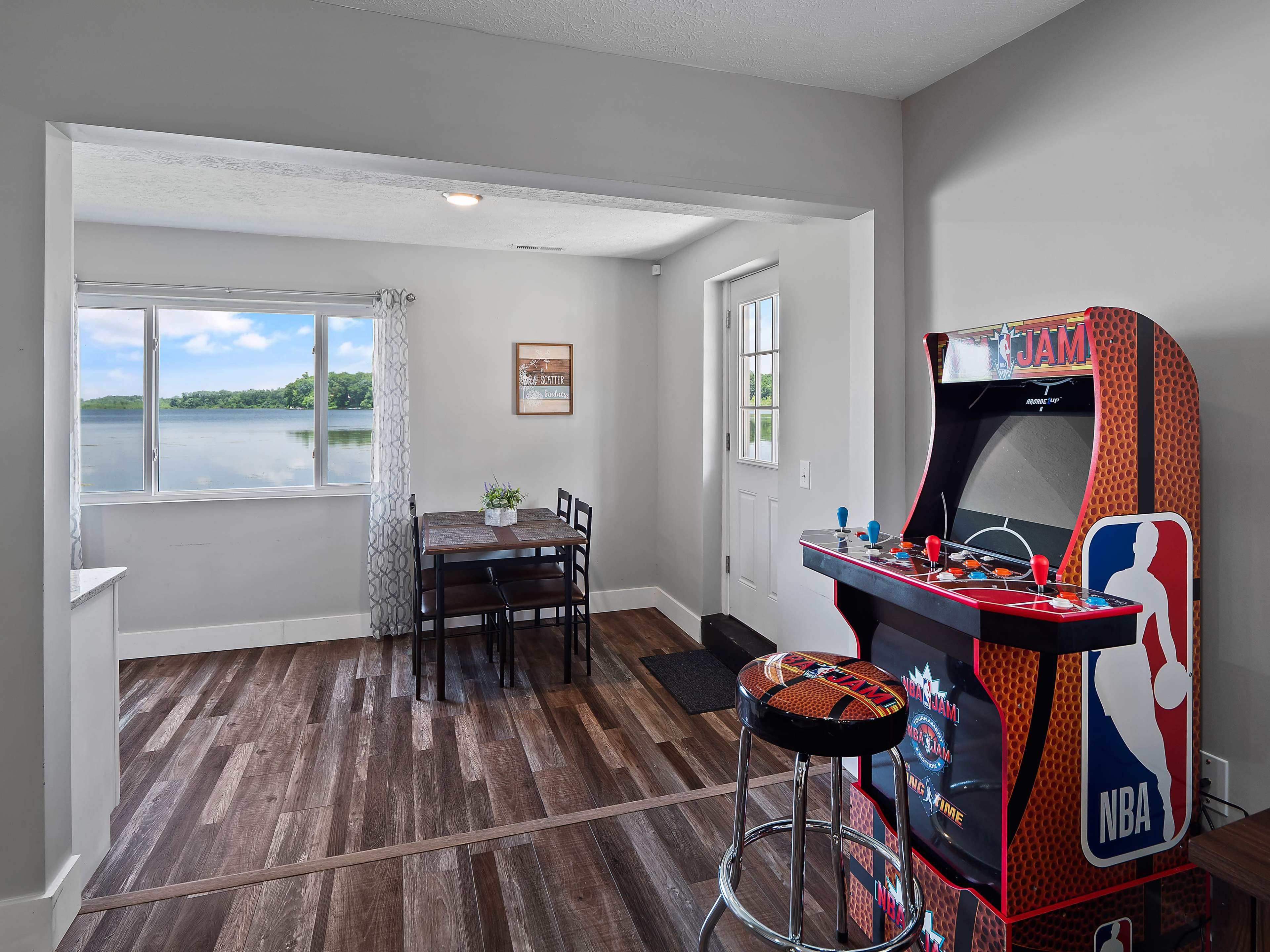 A game arcade machine is positioned next to a dining table in a room with a view of a lake through a large window.
