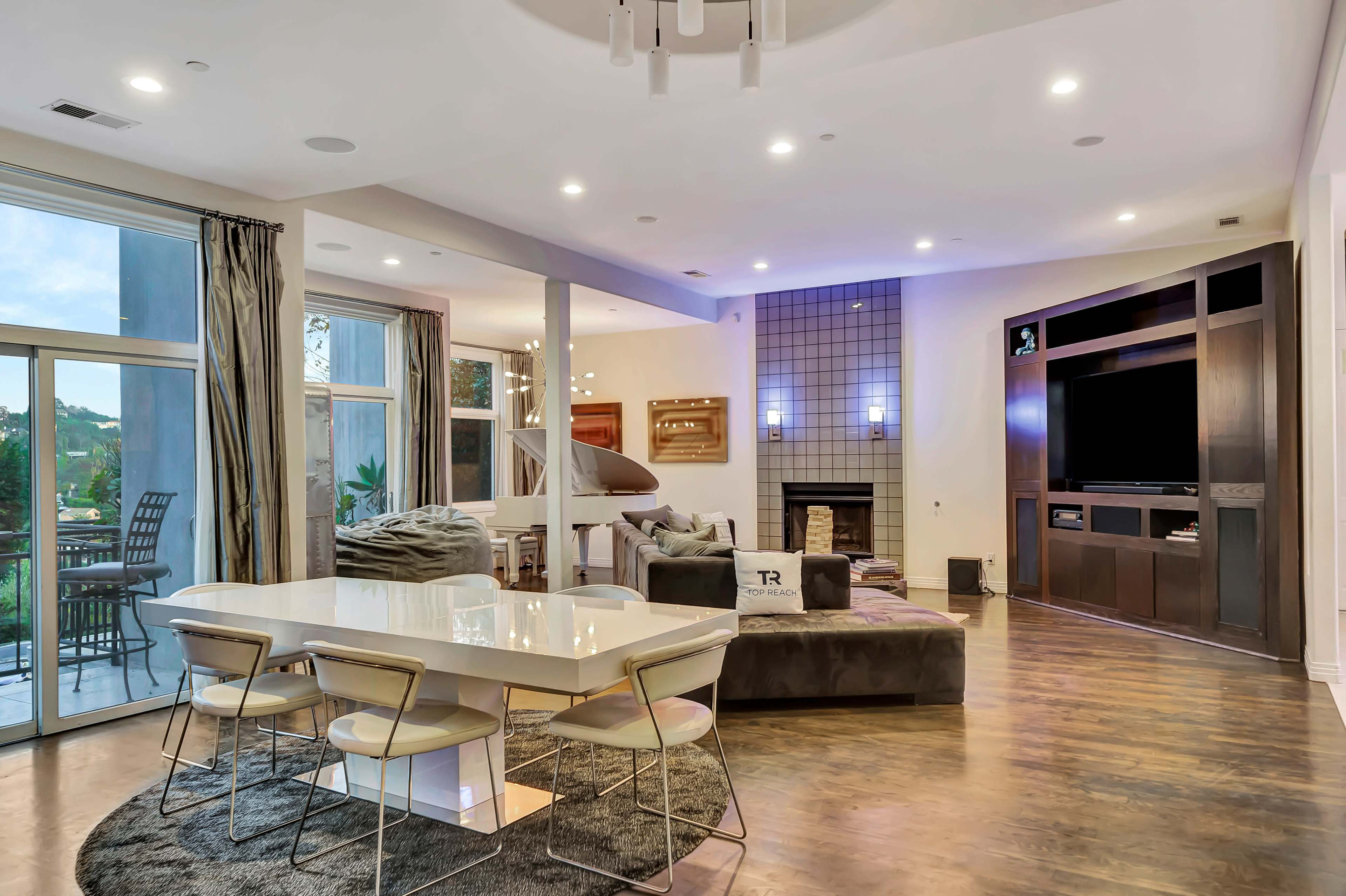 A spacious modern living area with a dining table, a large television unit, a fireplace, and large windows that provide outdoor views.