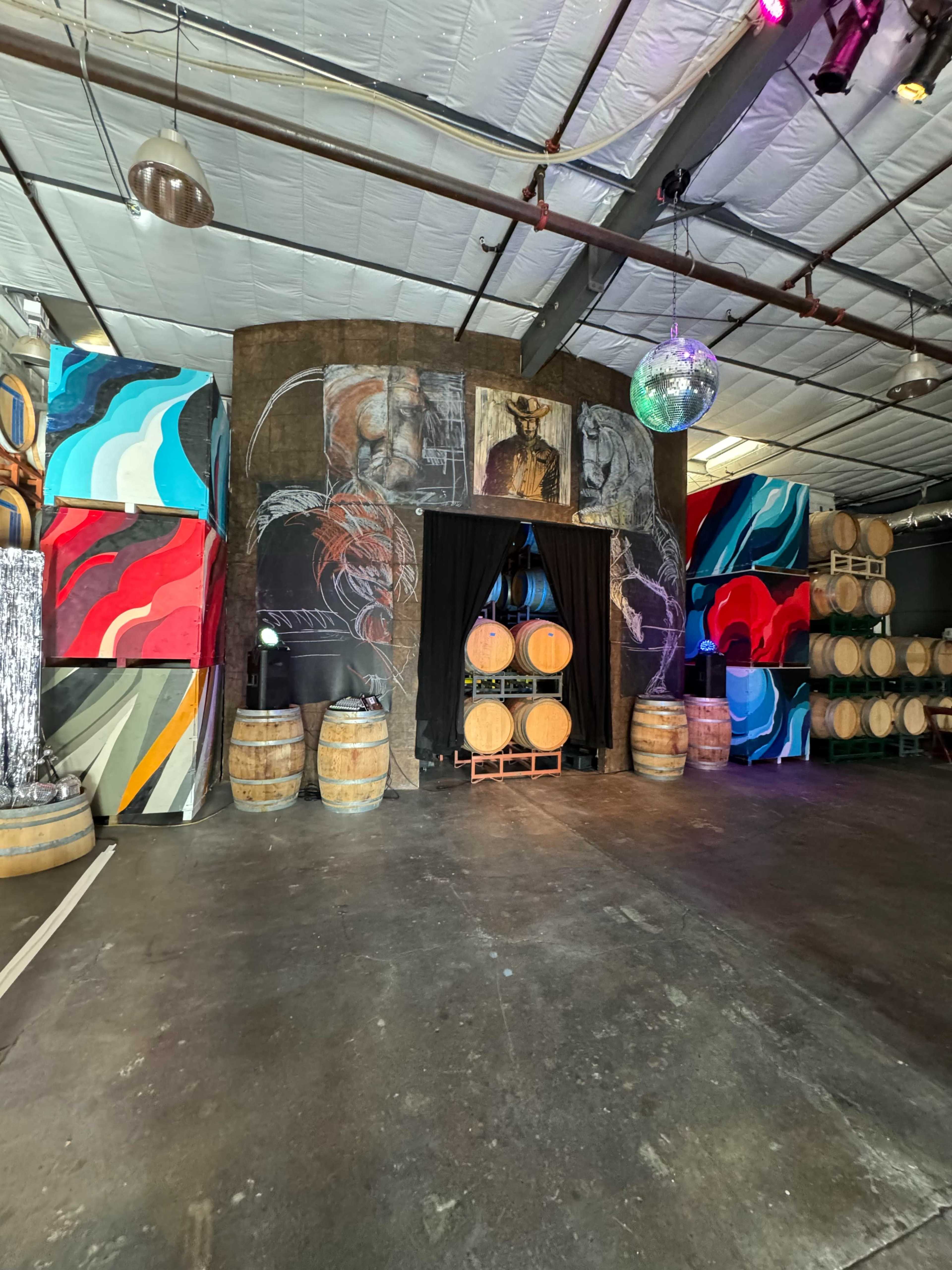 The interior of a large industrial space features colorful murals, wooden barrels arranged in stacks, and a black curtain at the center.