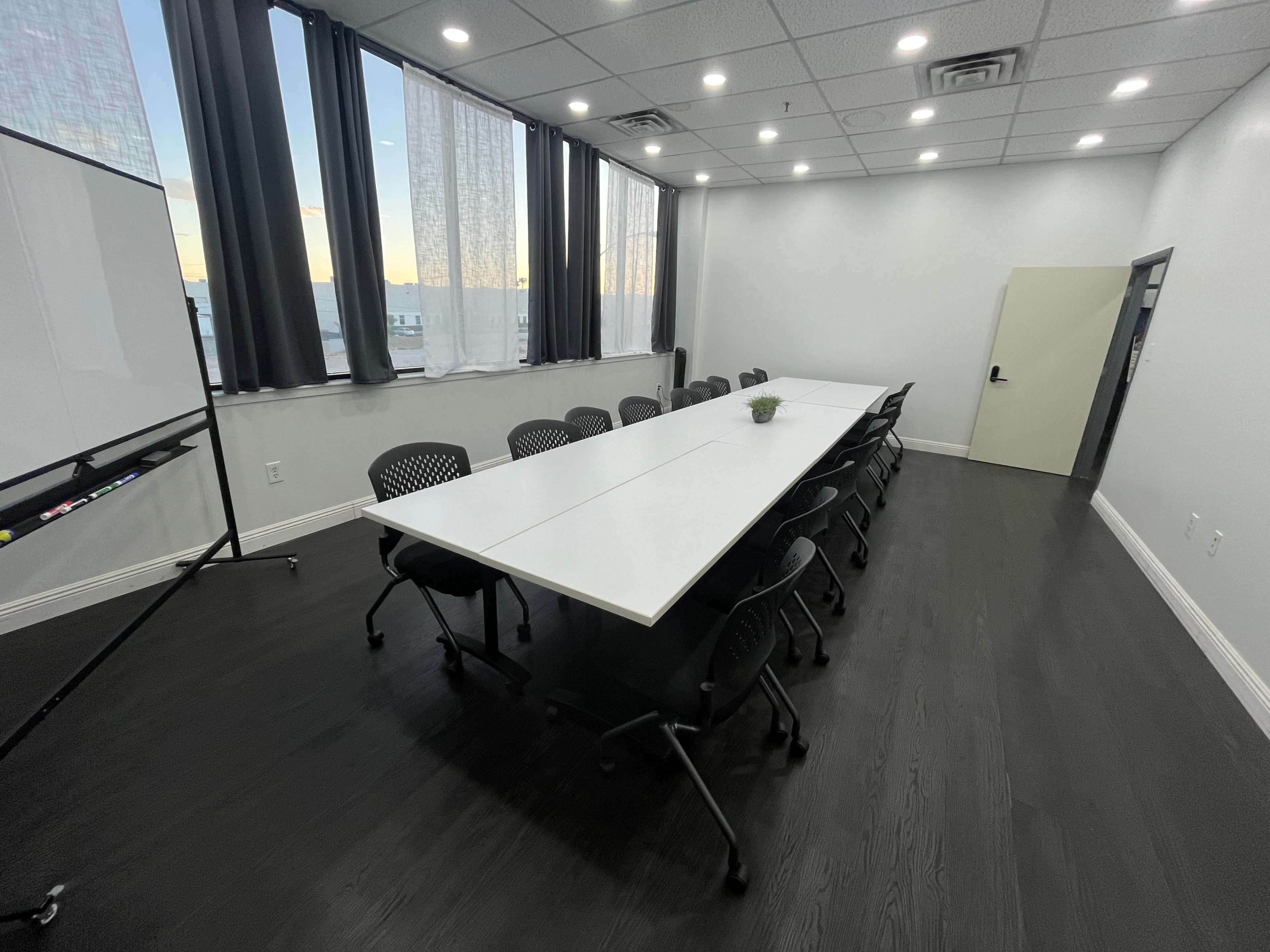 The image shows a modern conference room with a long white table surrounded by black chairs, large windows letting in natural light, and a whiteboard at one end.
