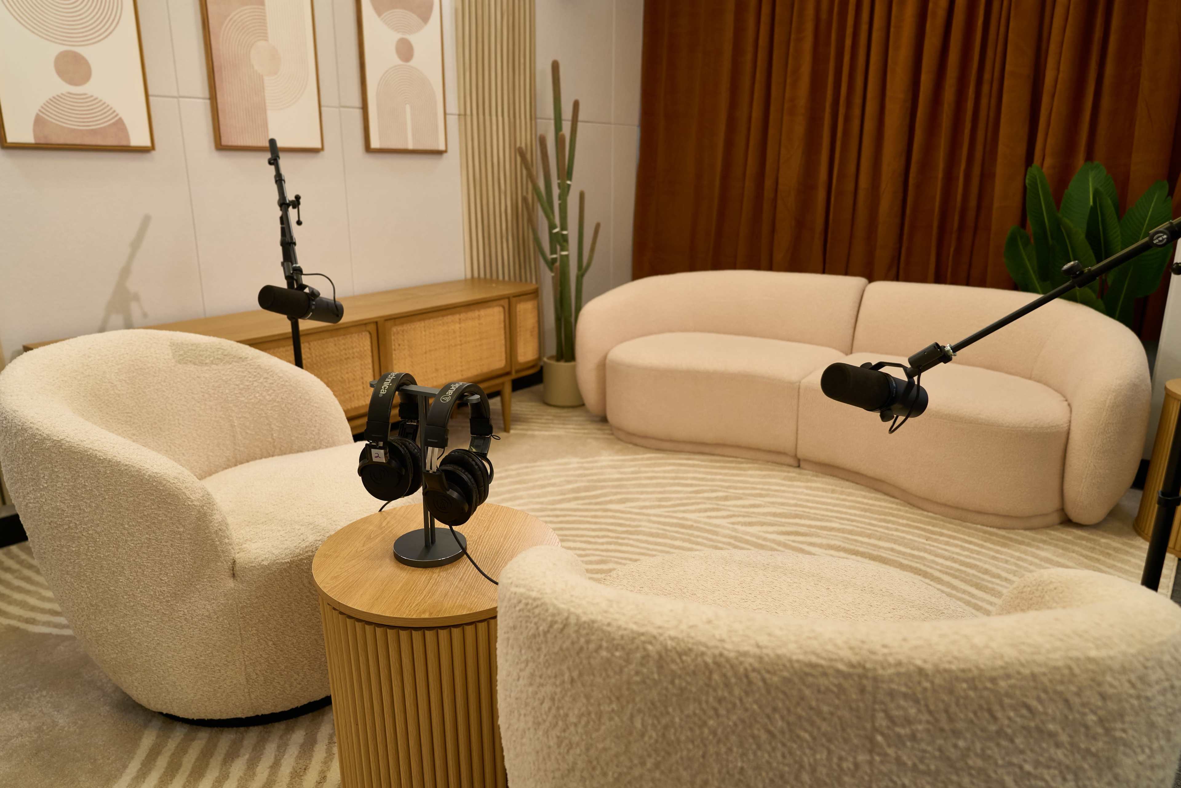 The image shows a comfortable recording studio setup with two plush armchairs, a small wooden table, and microphones mounted on stands, surrounded by decorative elements like plants and abstract art on the walls.