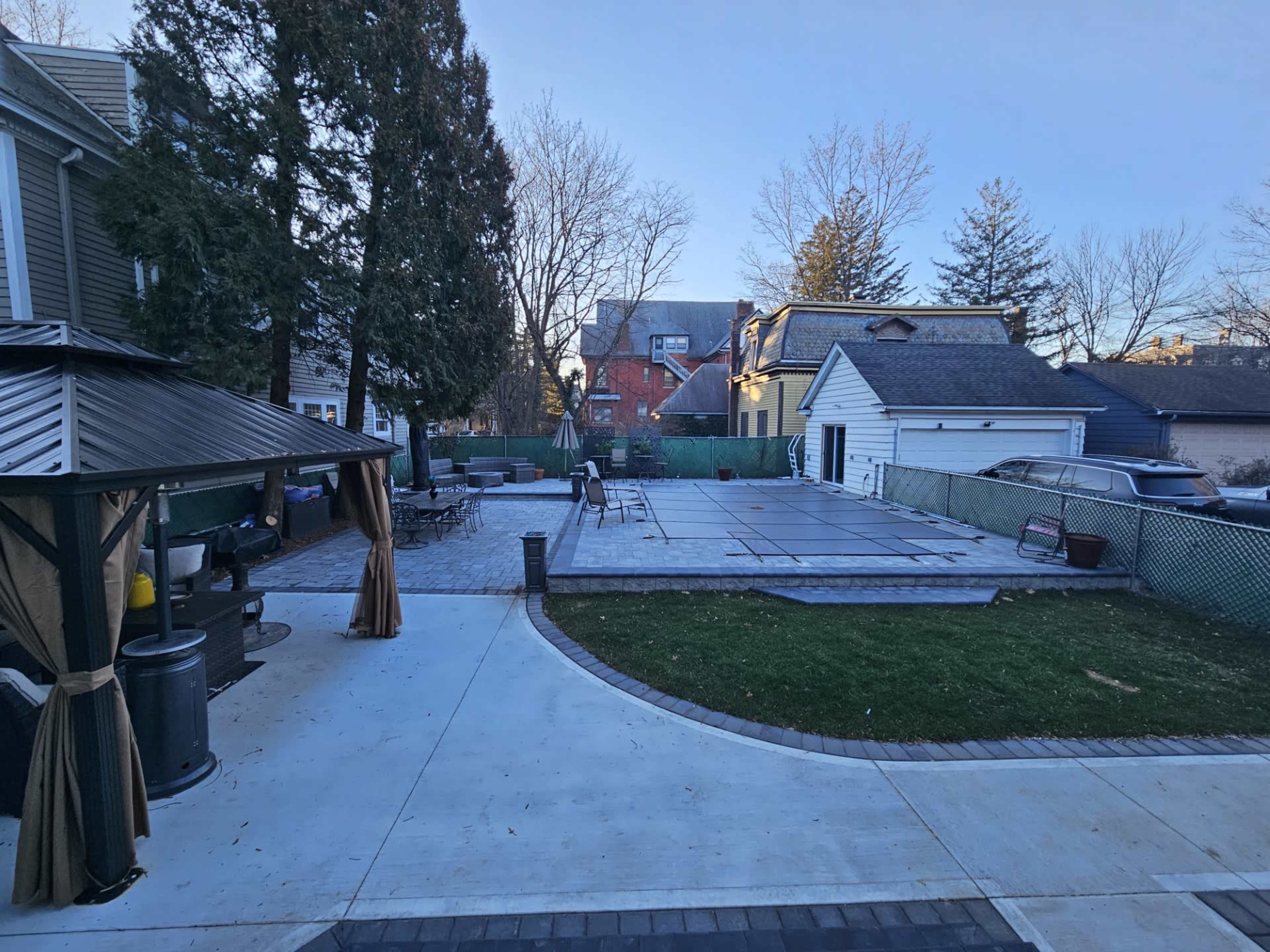A backyard with a covered seating area, a pool covered with a safety cover, and a paved patio area adjacent to a house.