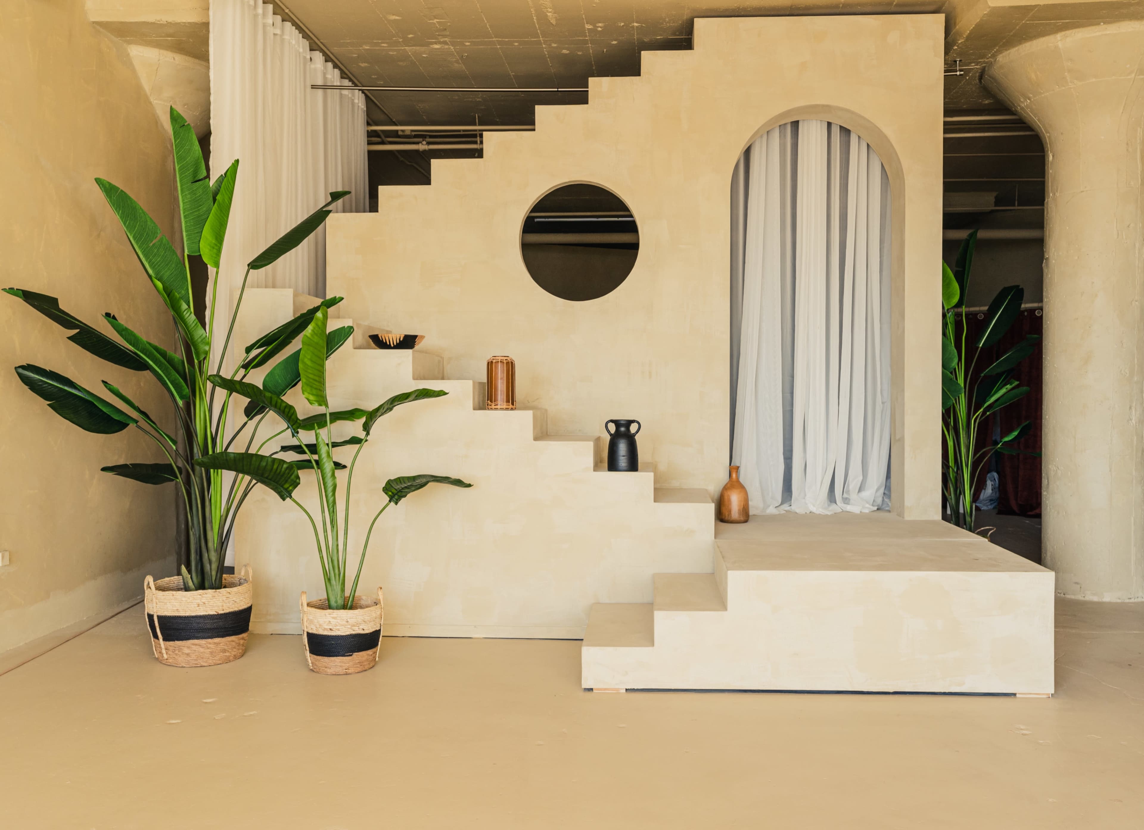 The image features a minimalist interior with a staircase leading to a raised platform, surrounded by large potted plants and a round mirror on the wall.