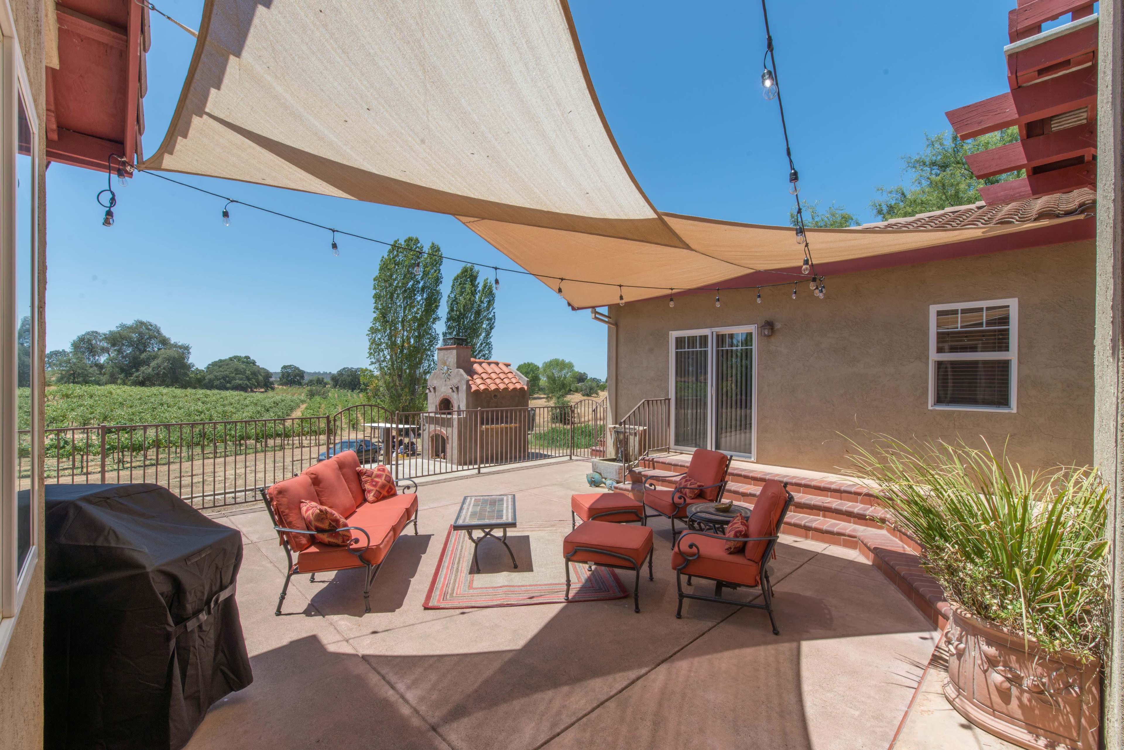 The image shows a patio area featuring red cushioned furniture, a barbecue grill, and a view of surrounding greenery under a shade canopy.