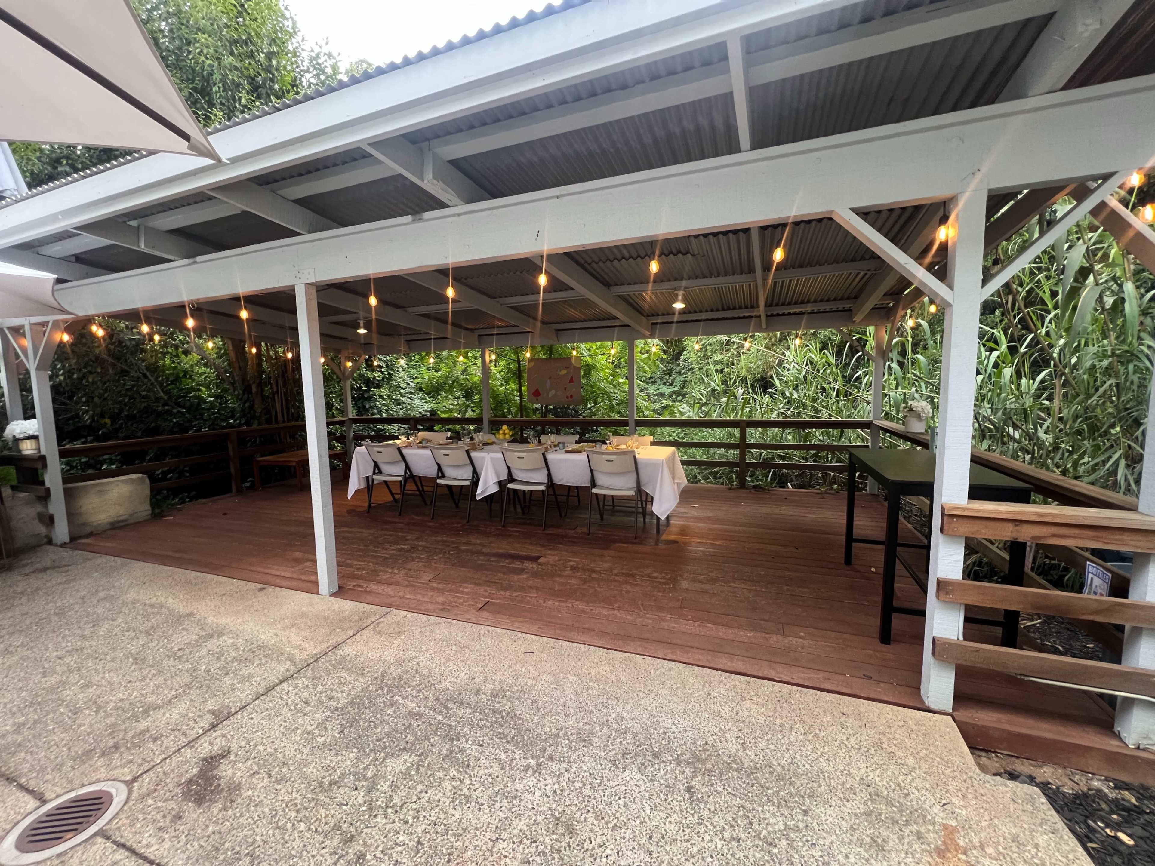 A covered outdoor dining space features a long table set with chairs, surrounded by greenery and illuminated by string lights.