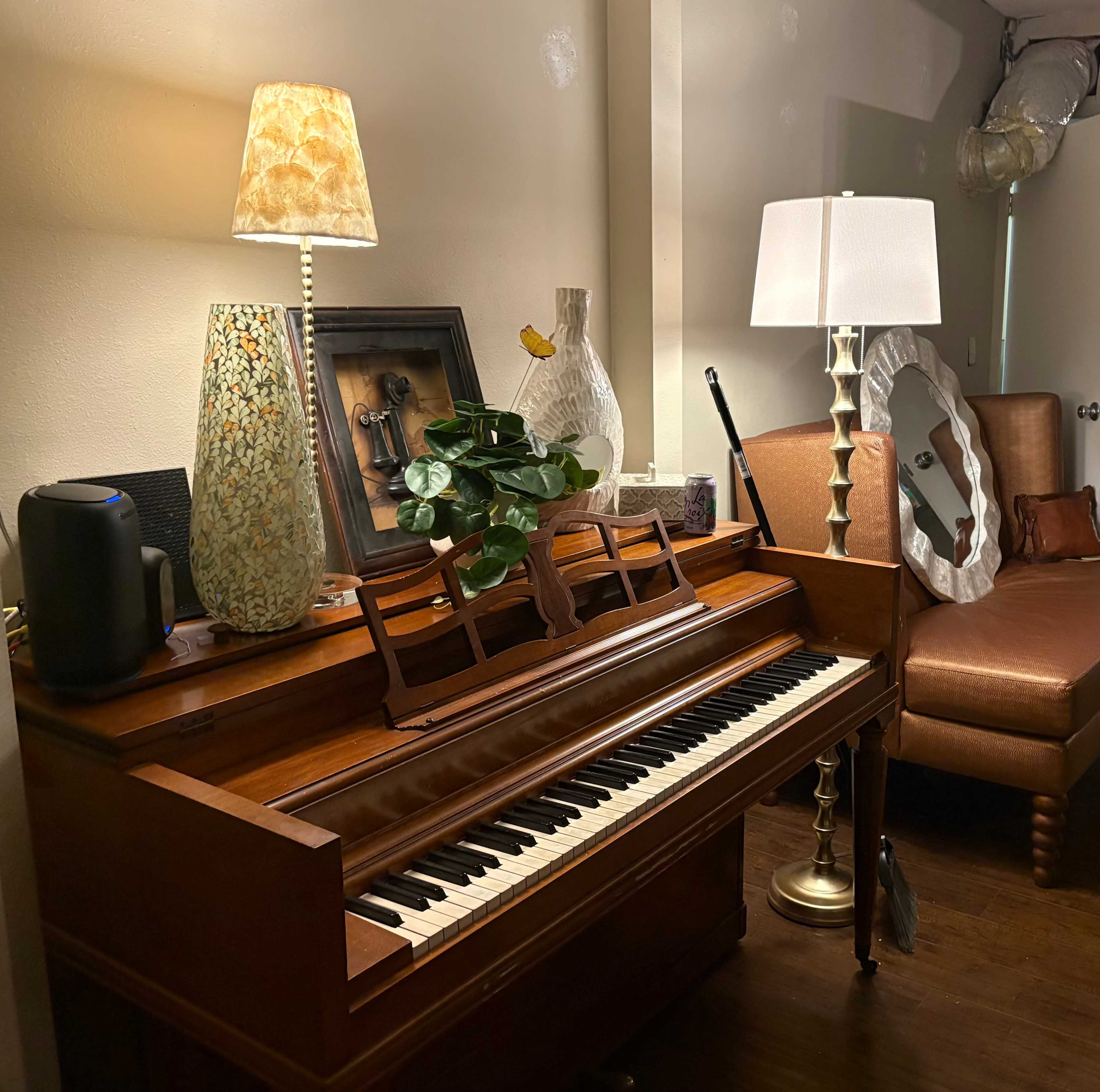 A wooden piano is positioned beside a lamp, a decorative vase, and a plant, with a couch visible in the background.