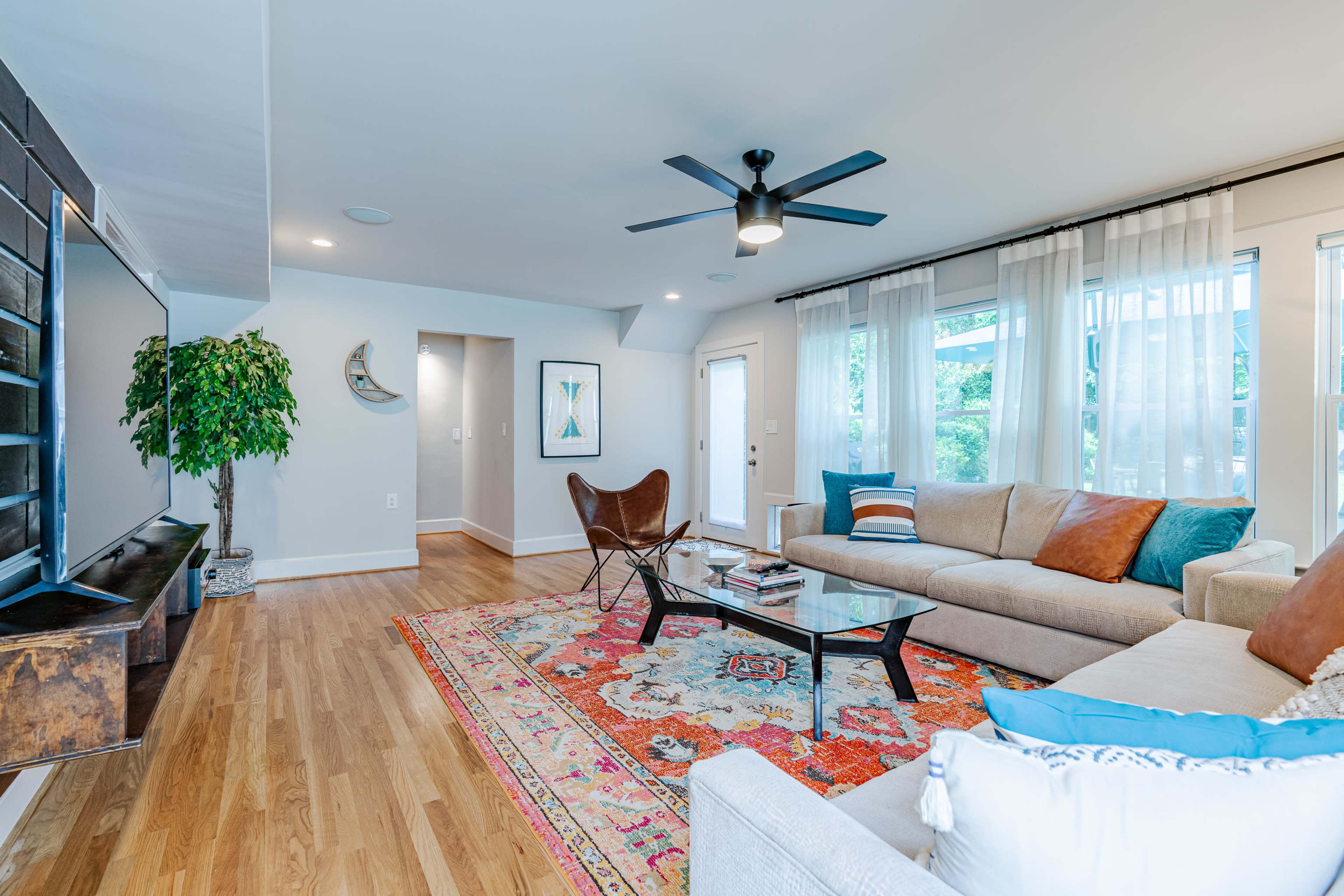 A modern living room features a sectional sofa, a butterfly chair, a glass coffee table, and a colorful area rug, with large windows allowing natural light to enter.