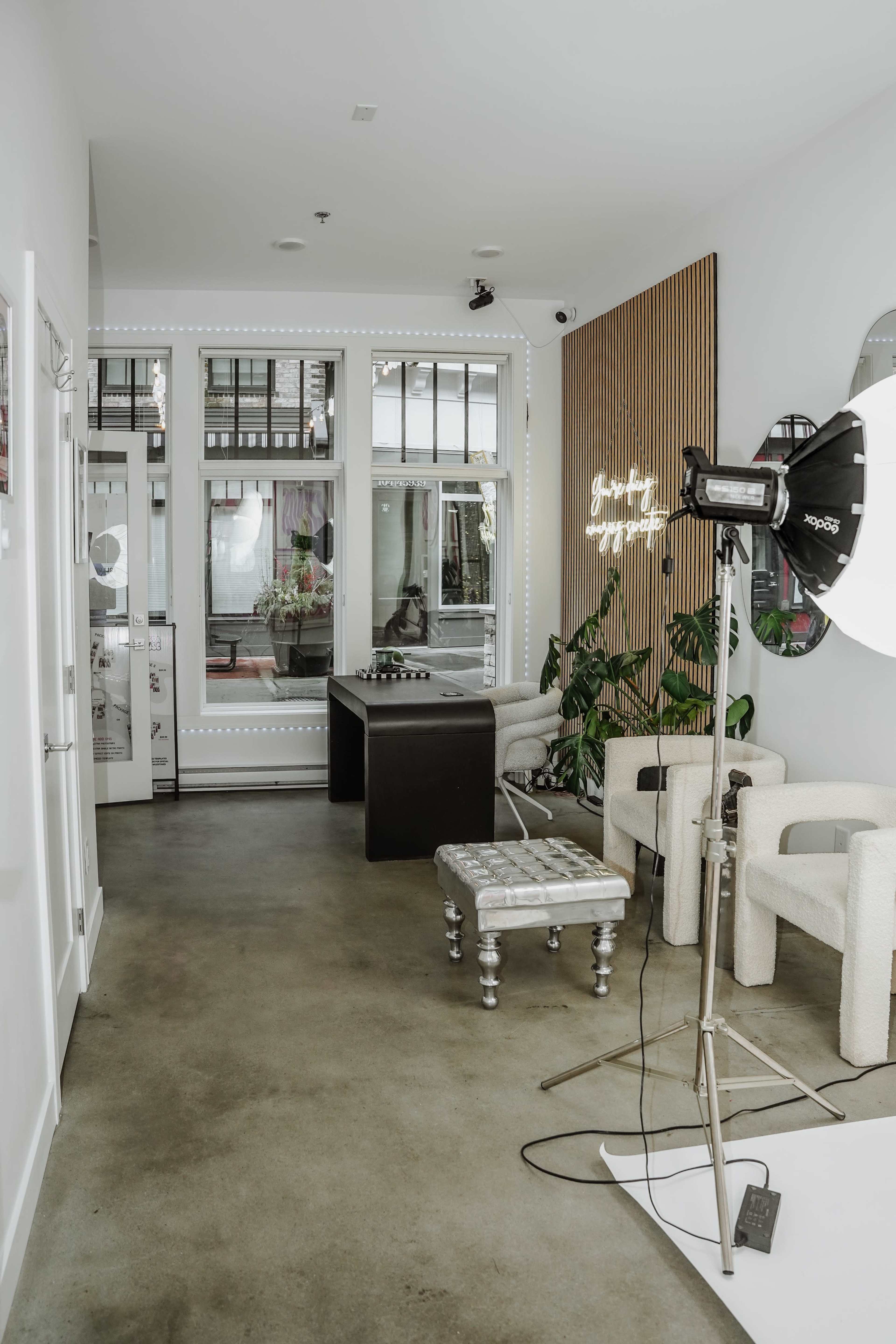 The image depicts a modern, minimalist interior of a salon or studio featuring a seating area, a light setup, and large windows allowing natural light.