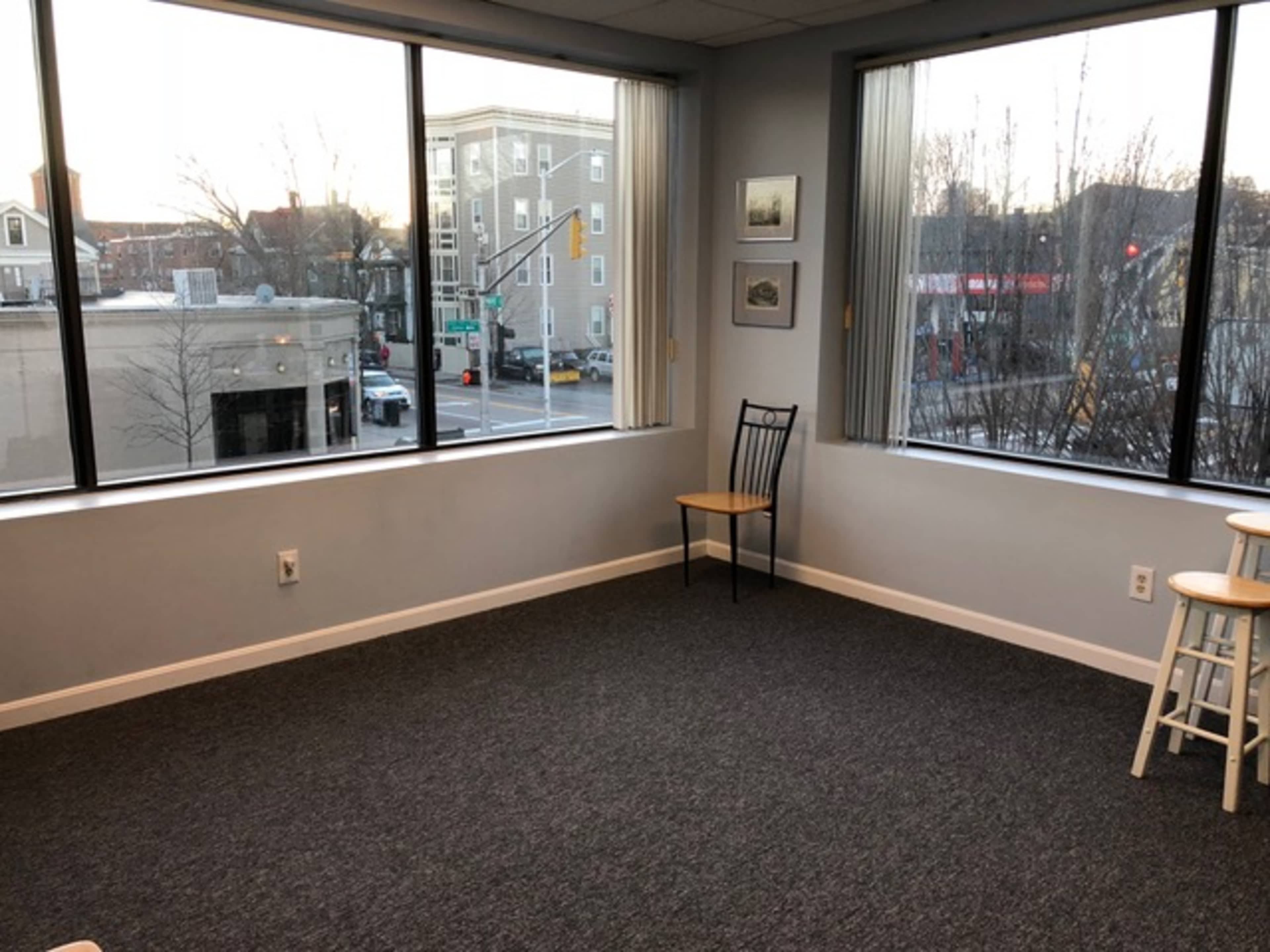 A vacant room with two large windows overlooking a street and a few pieces of furniture, including a black chair and two stools.