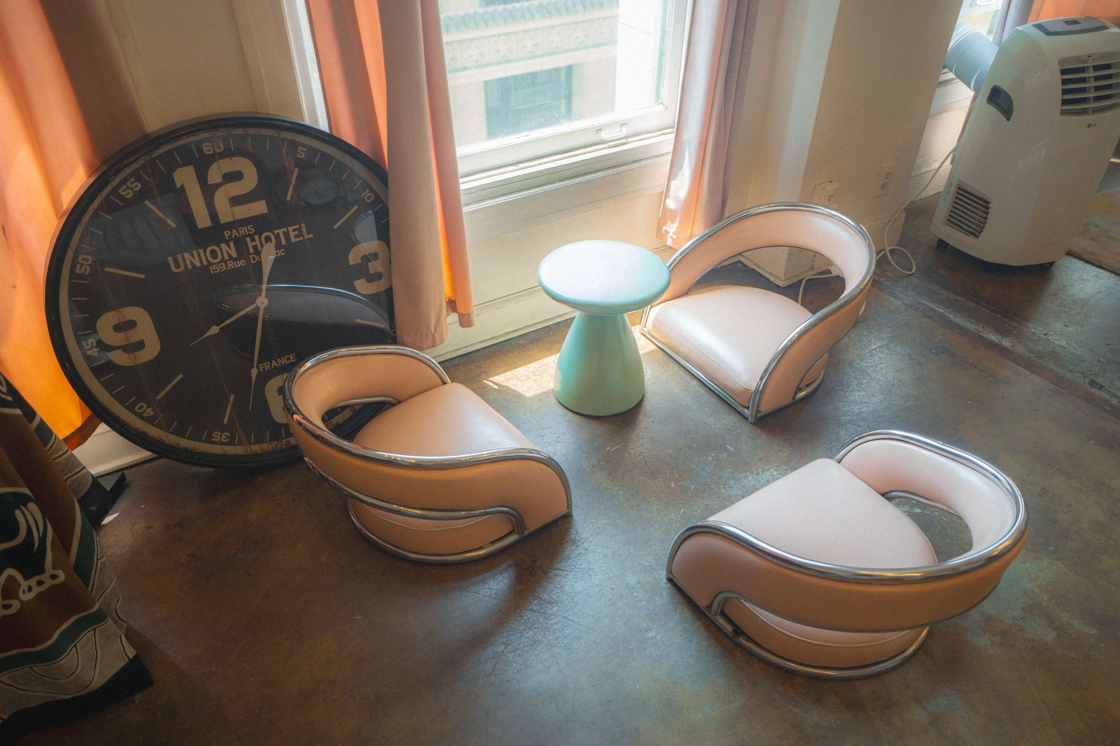 The image shows a room featuring three pink and silver chairs arranged around a small green table, with a large wall clock displaying the time nearby.