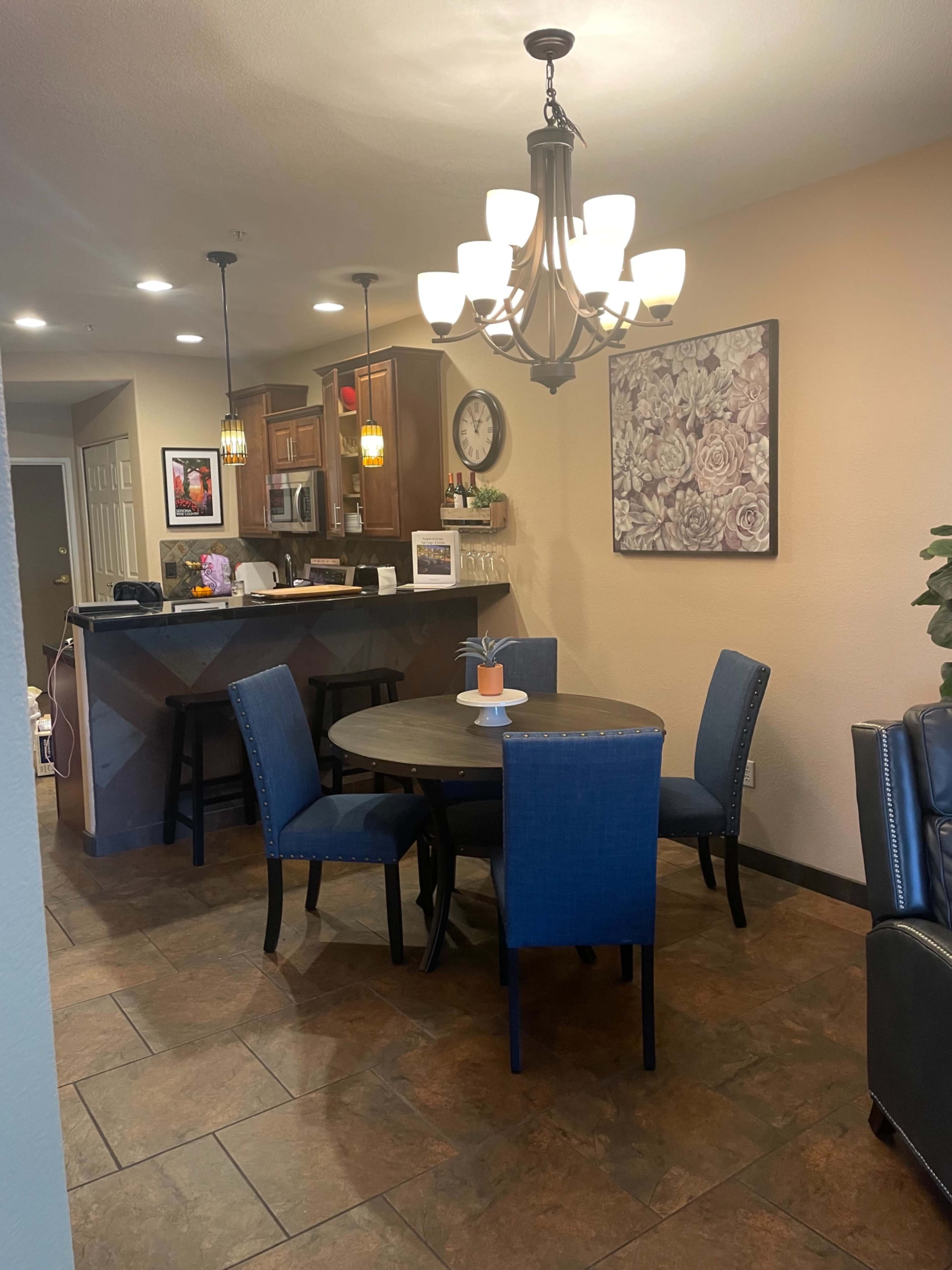 The image shows a dining area with a round table and four blue chairs, adjacent to a kitchen and inviting living space.