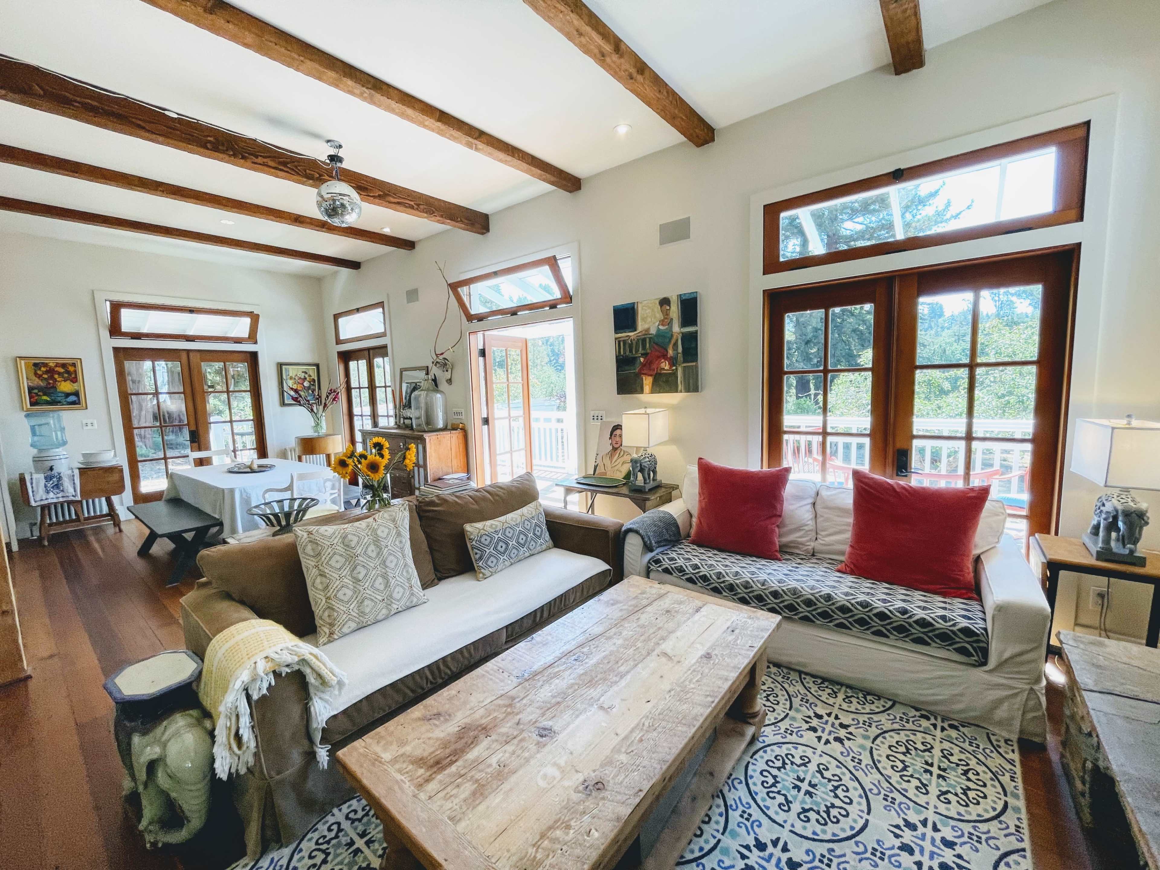 A bright living room features wooden beams, large windows, a rustic wooden coffee table, and various pieces of furniture arranged around a patterned rug.