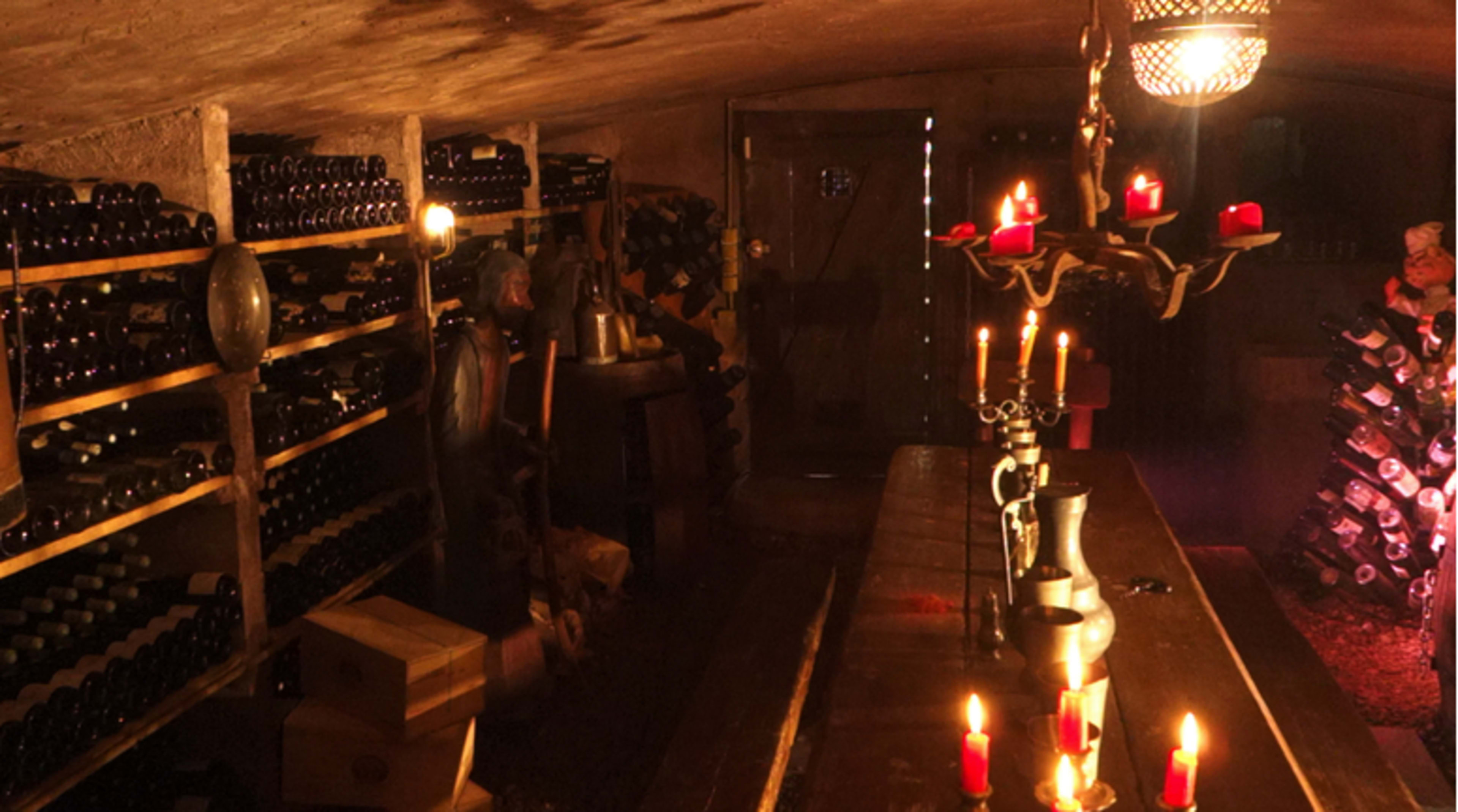 A dimly lit wine cellar with wooden shelves filled with bottles, a table at the center, and a chandelier with candles providing illumination.