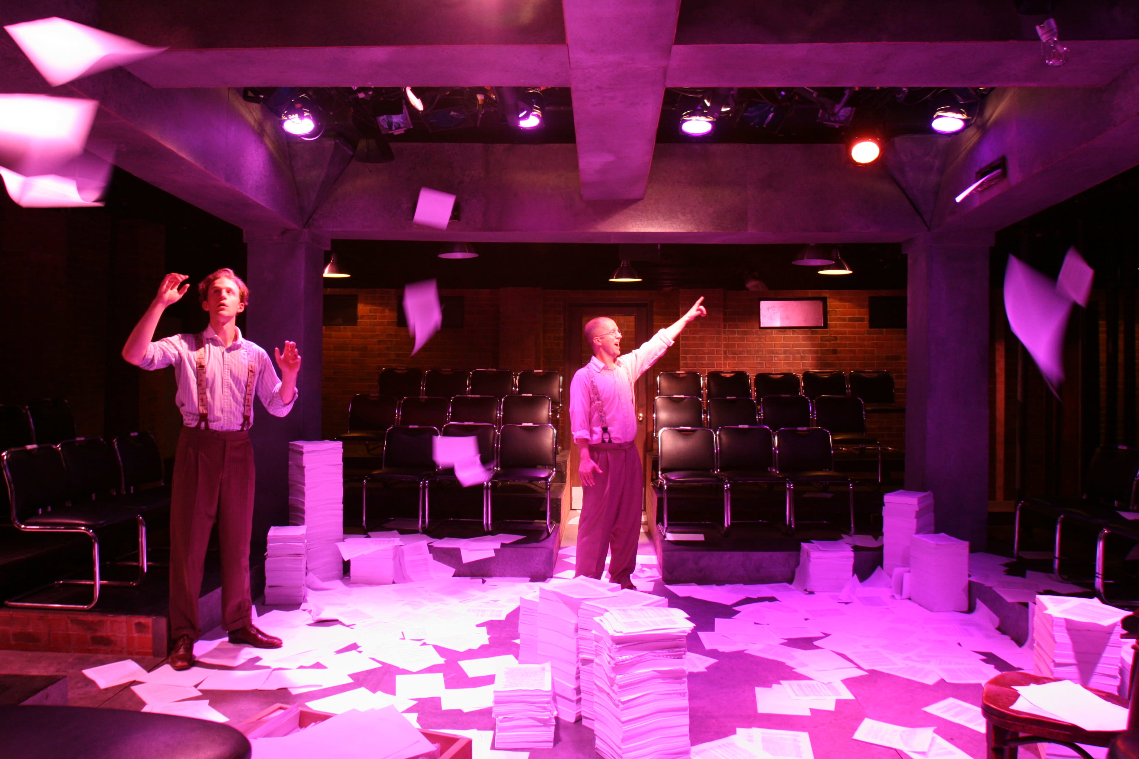 Two men in suspenders are standing in a theater space surrounded by scattered papers, with lights illuminating the scene from above.