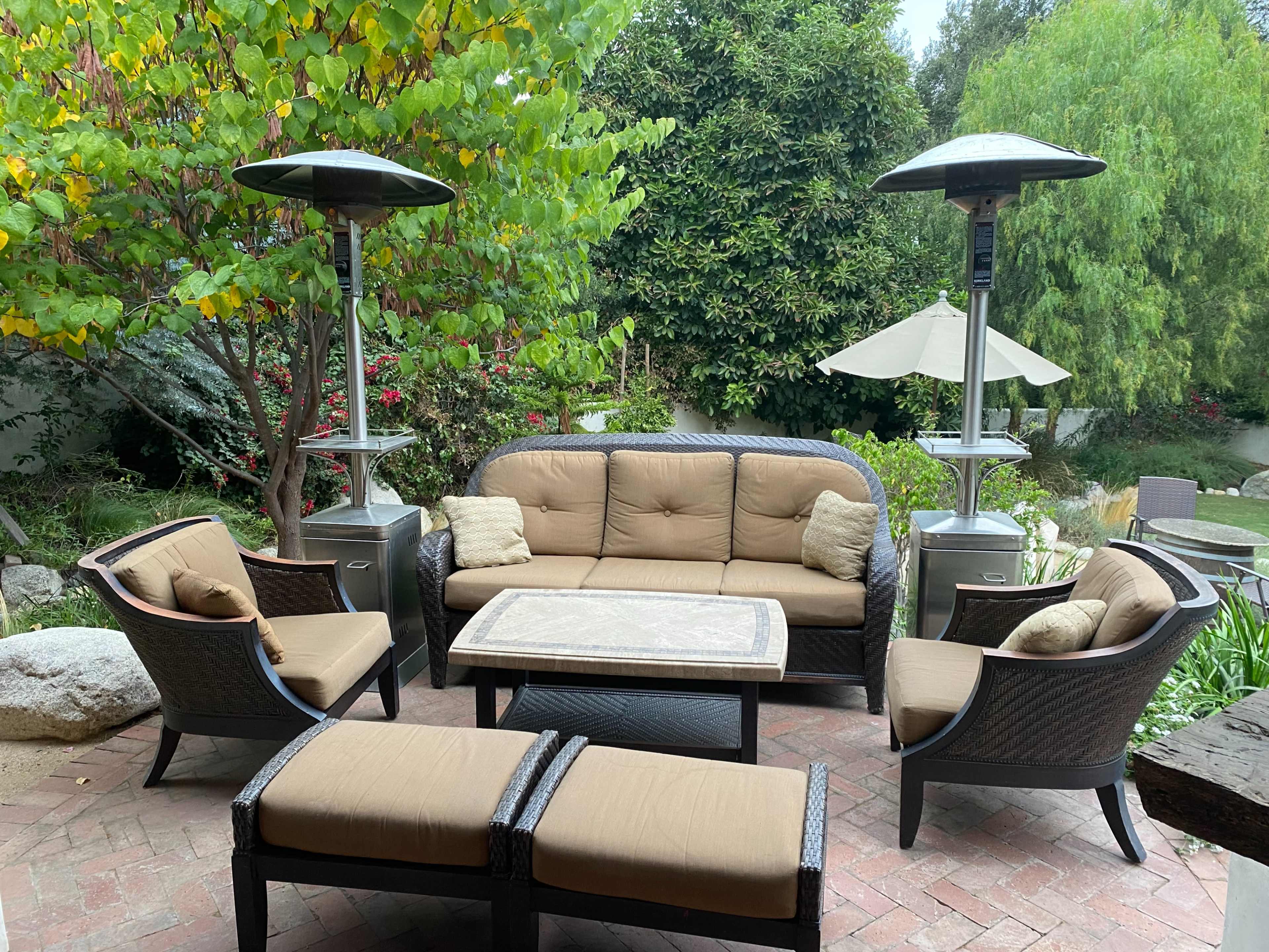 A patio seating area features cushioned furniture arranged around a rectangular table, with heating lamps and greenery in the background.