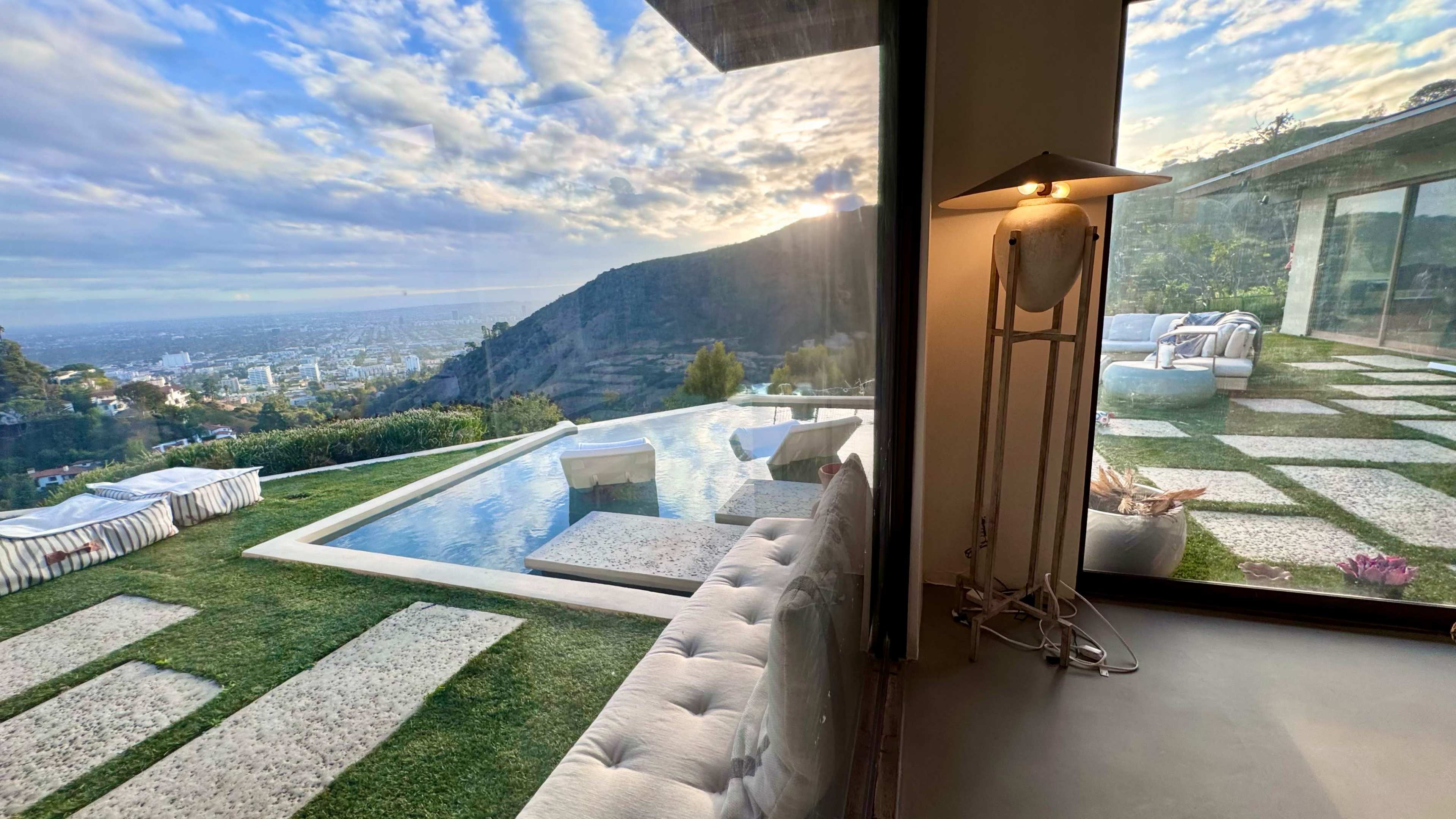The image shows a modern interior with a large window overlooking a hillside swimming pool, surrounded by landscaped greenery and a city skyline in the distance.
