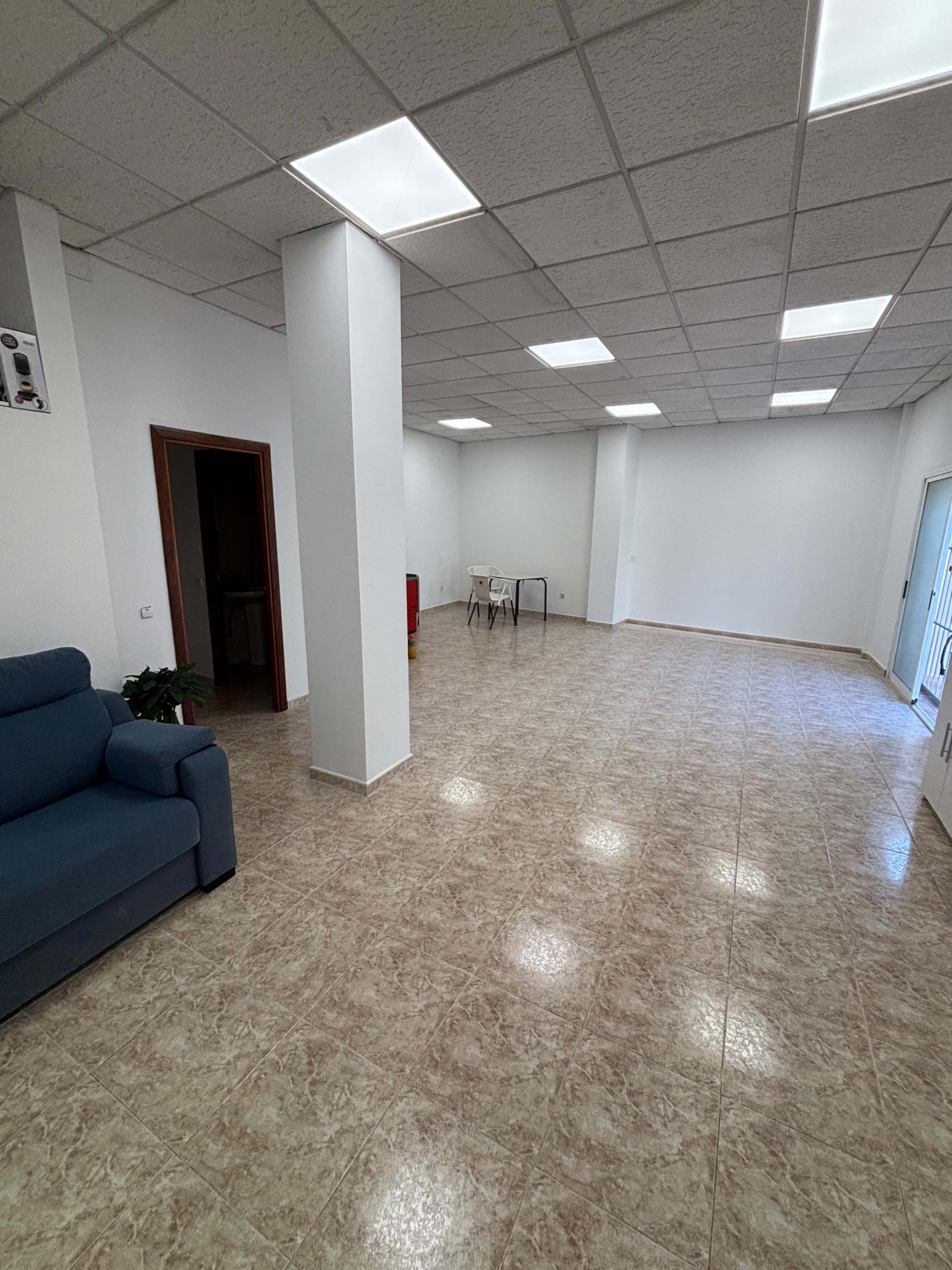 The image shows a spacious, empty room with light-colored tiled flooring, white walls, and a few pieces of furniture, including a blue couch and a table with chairs.