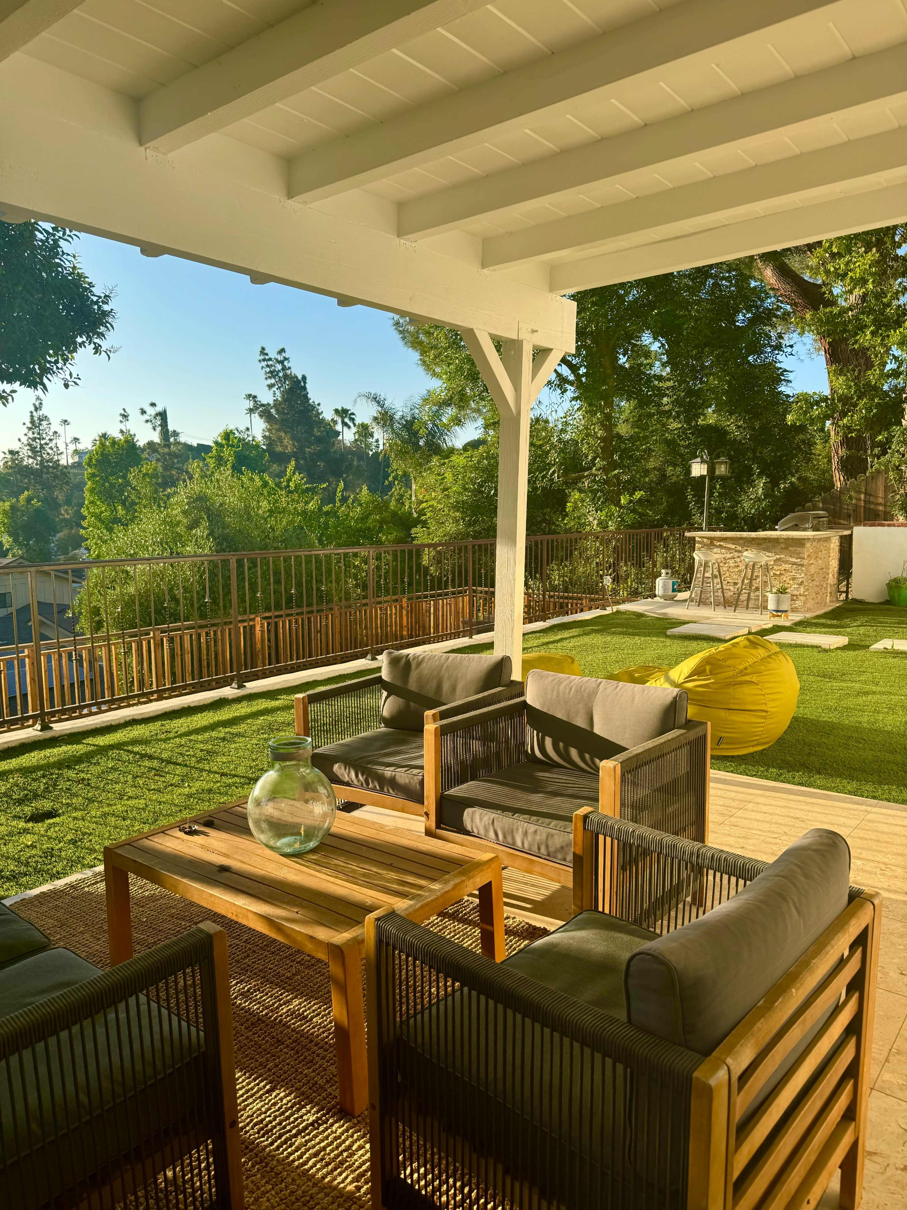 The image shows an outdoor living space with a wooden table and seating arranged under a covered patio, surrounded by greenery and a well-maintained lawn.