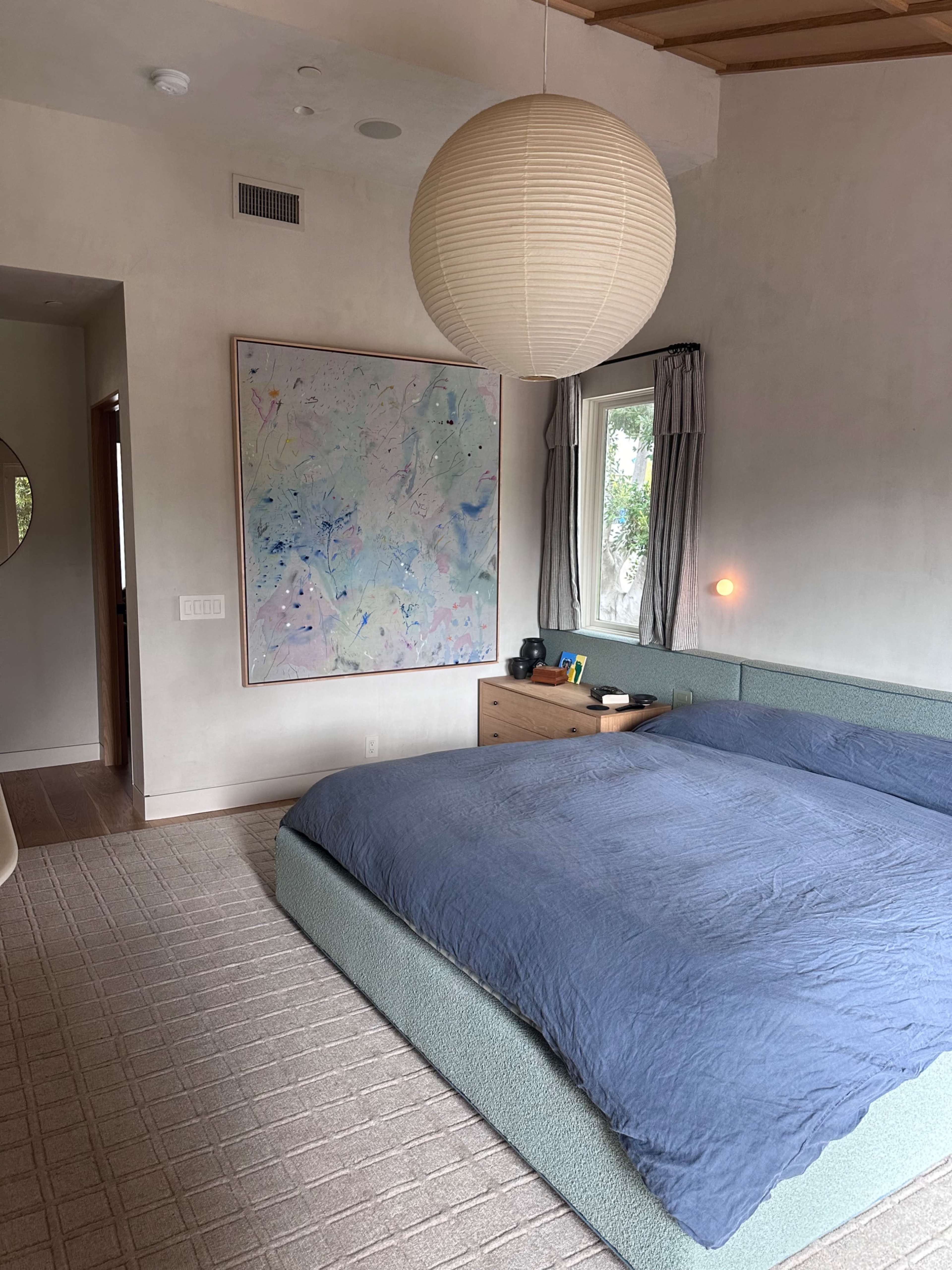A modern bedroom featuring a large bed with a blue duvet, a piece of abstract art on the wall, and a round paper lantern hanging from the ceiling.