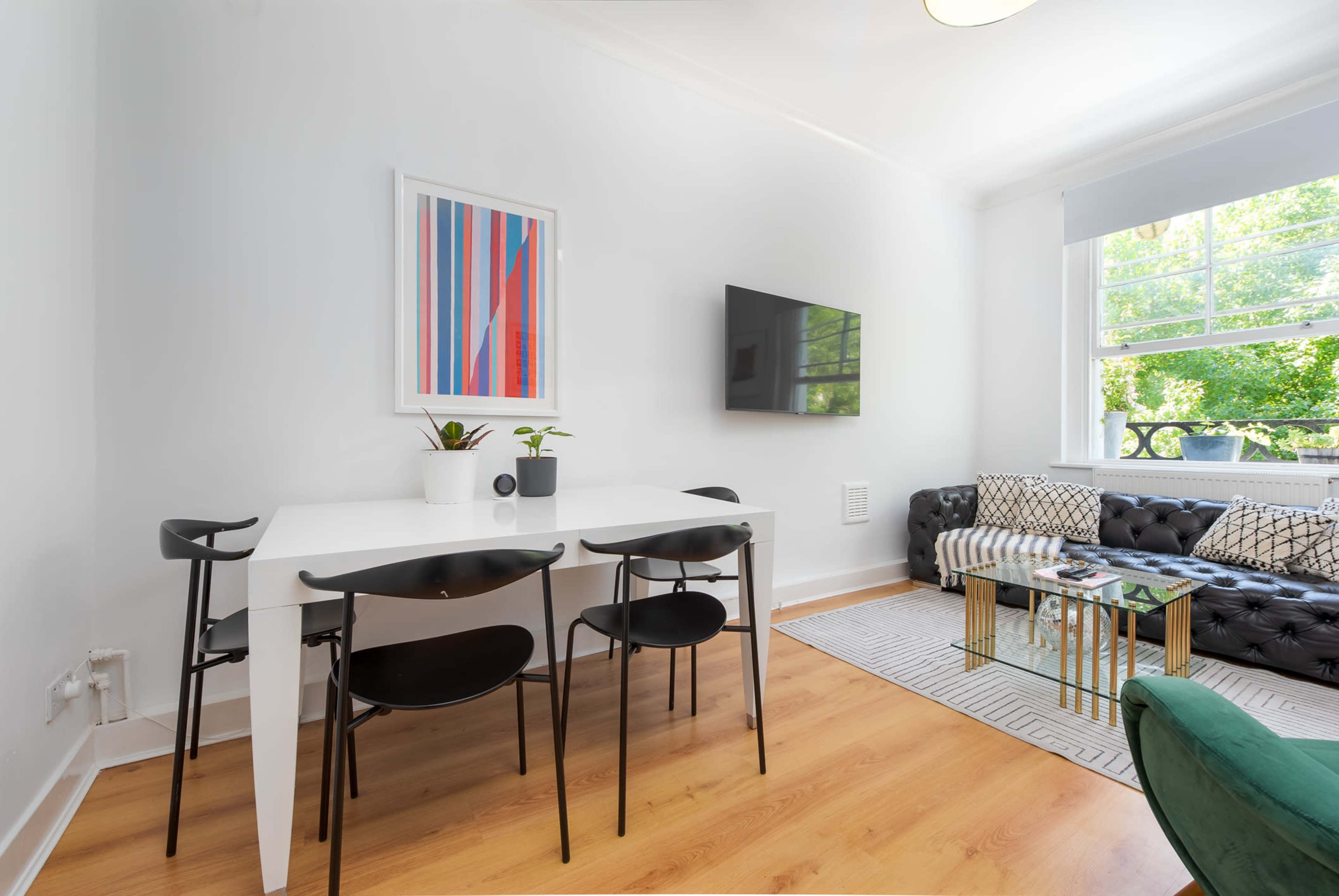A modern living space features a black and white dining table set, a green chair, a television mounted on the wall, and large windows letting in natural light.
