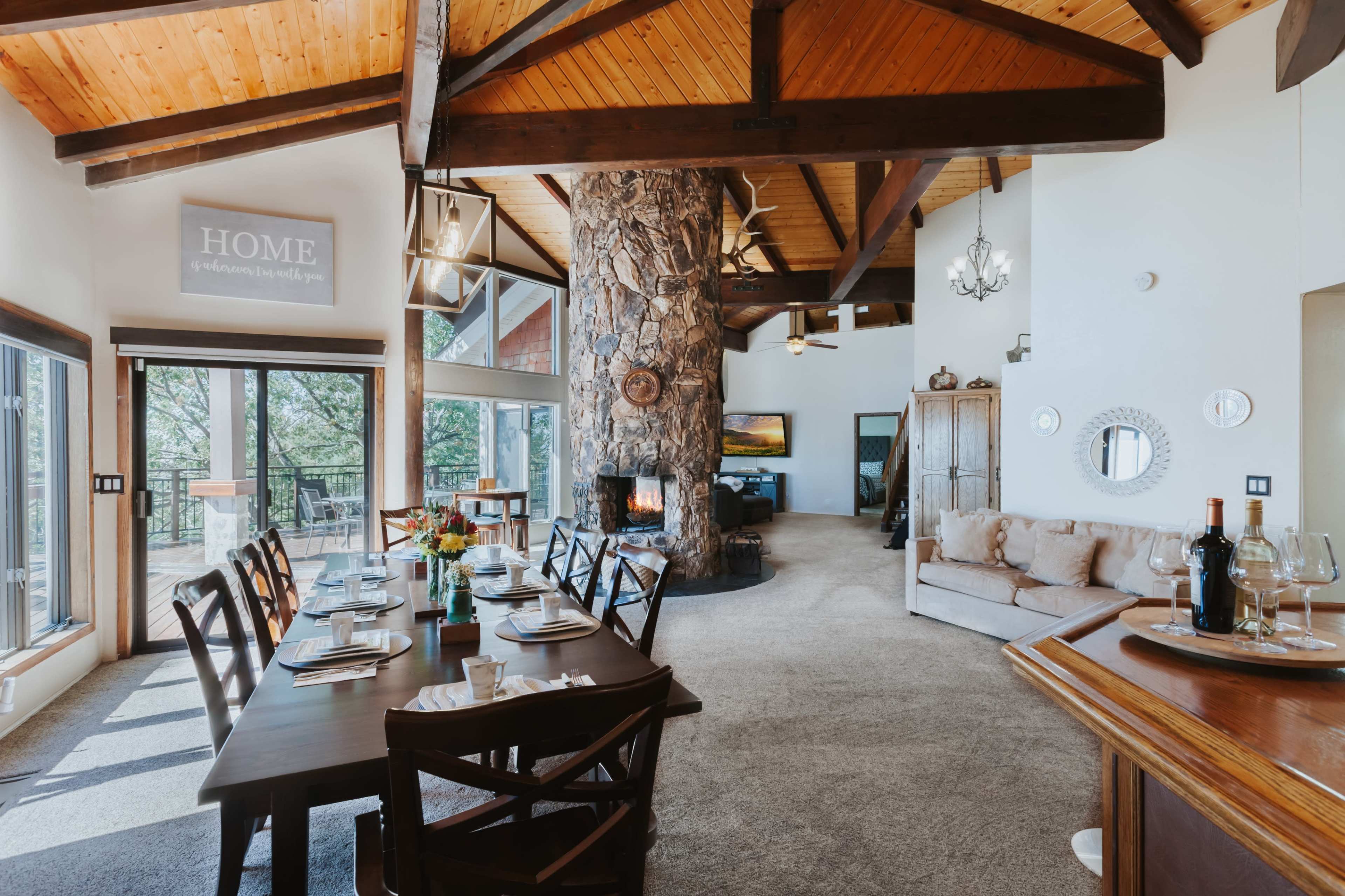 A spacious open-concept living and dining area featuring a large wooden table, a stone fireplace, and large windows with a view.