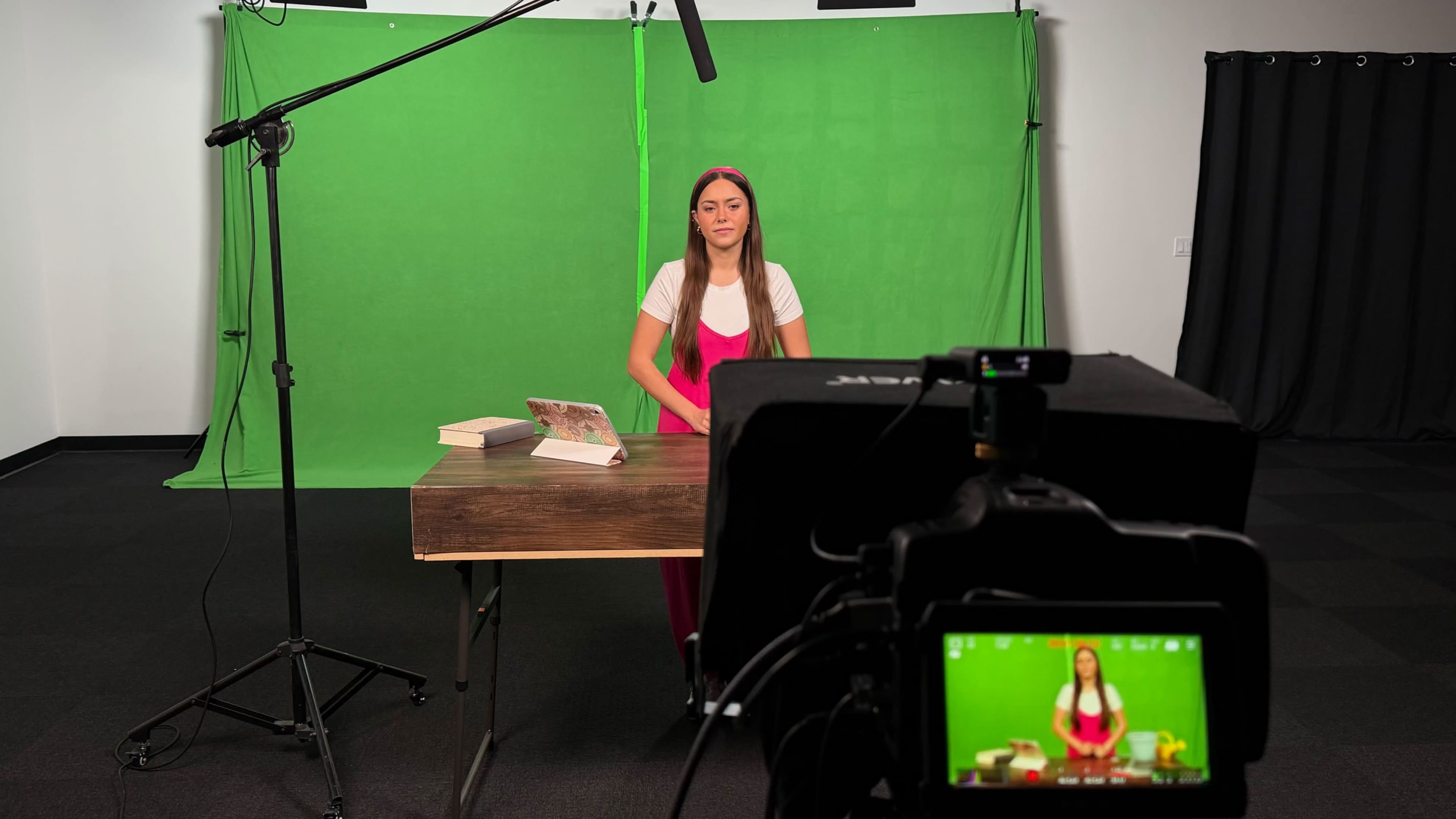 A woman in a pink outfit sits at a wooden table in front of a green screen with a camera focused on her.