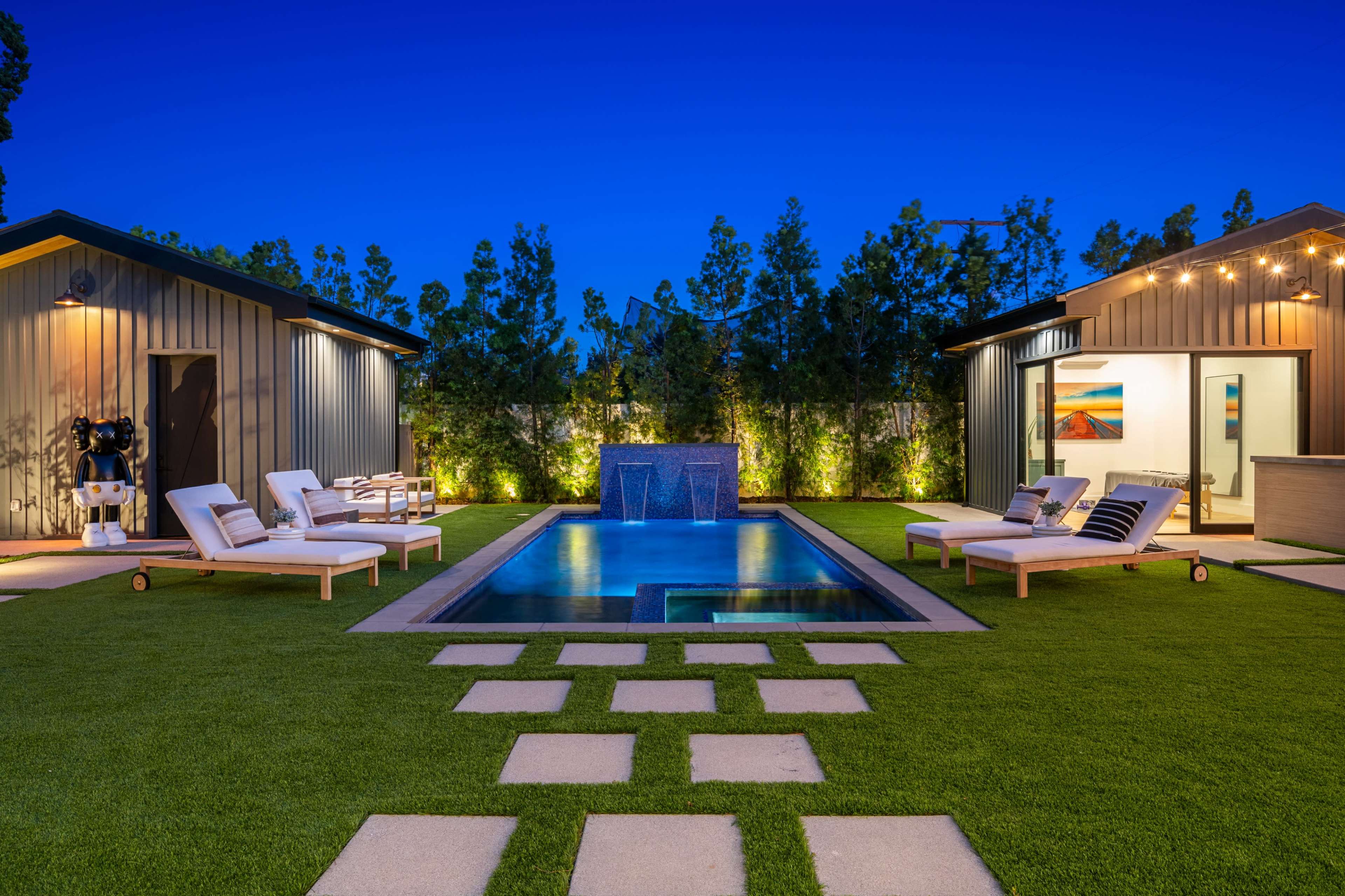 The image shows a modern backyard with a swimming pool framed by two separate structures, lounge chairs, and landscaped greenery at dusk.