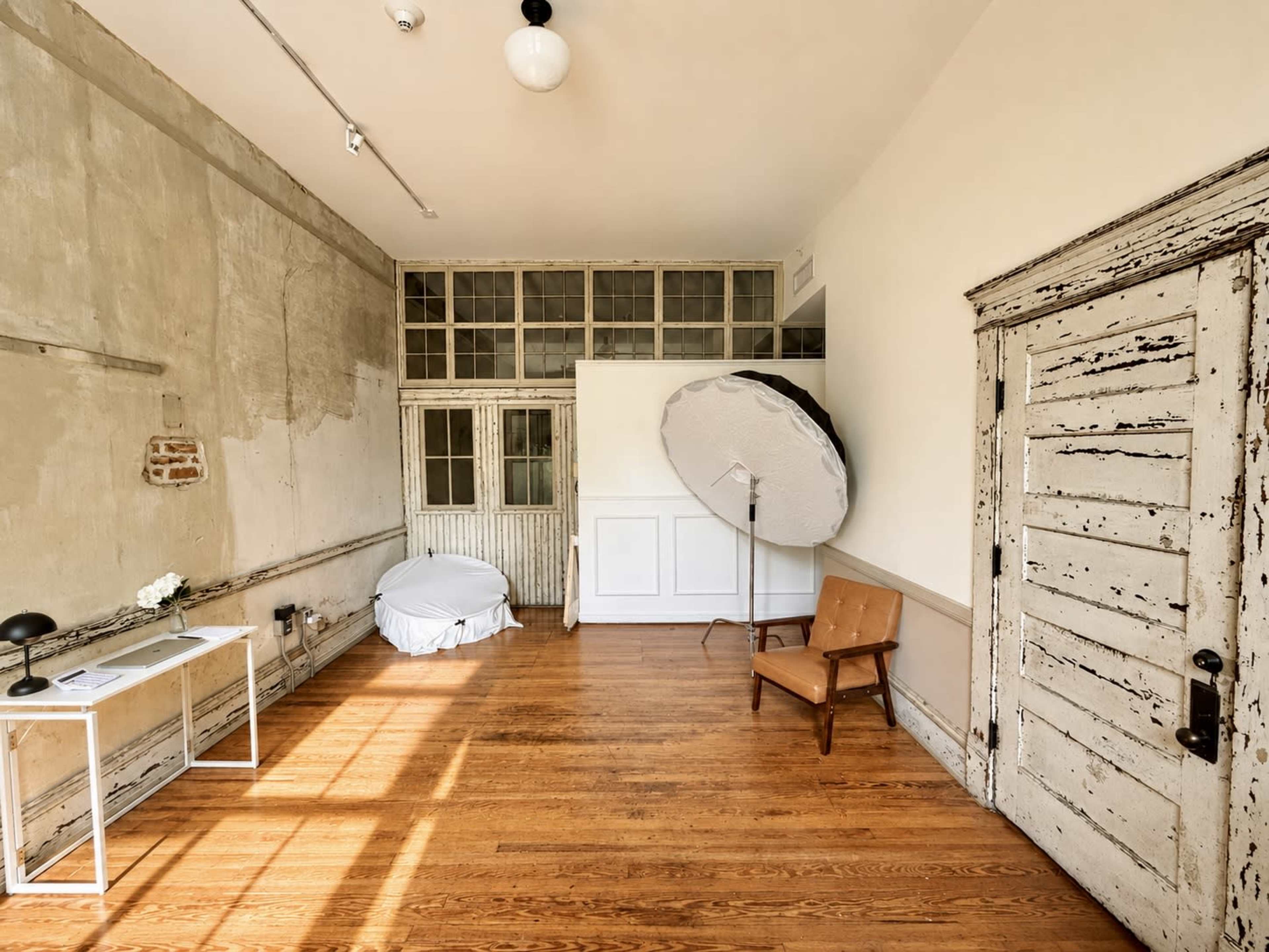 The image shows a spacious, well-lit studio with wooden floors, an old door, a white draped chair, a large round reflector, and a minimalist table.
