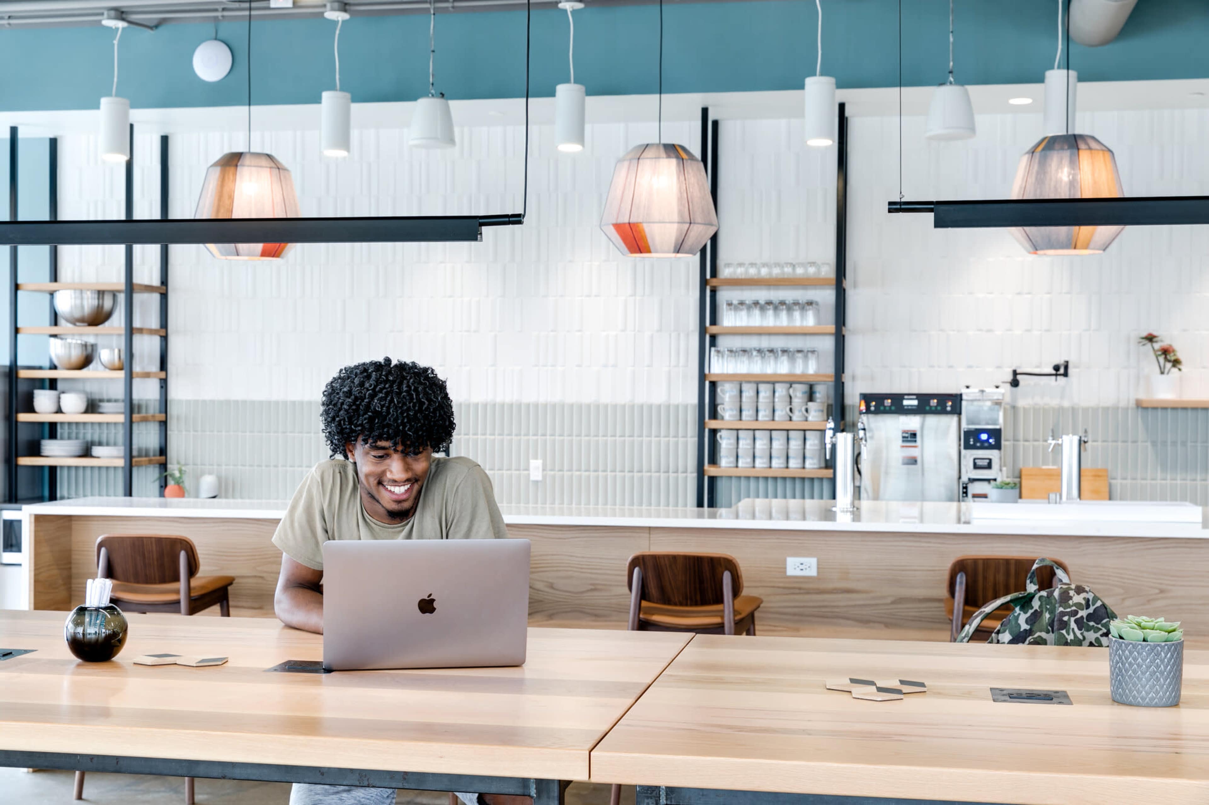 A young man with curly hair sits at a large wooden table in a modern café, working on a laptop.