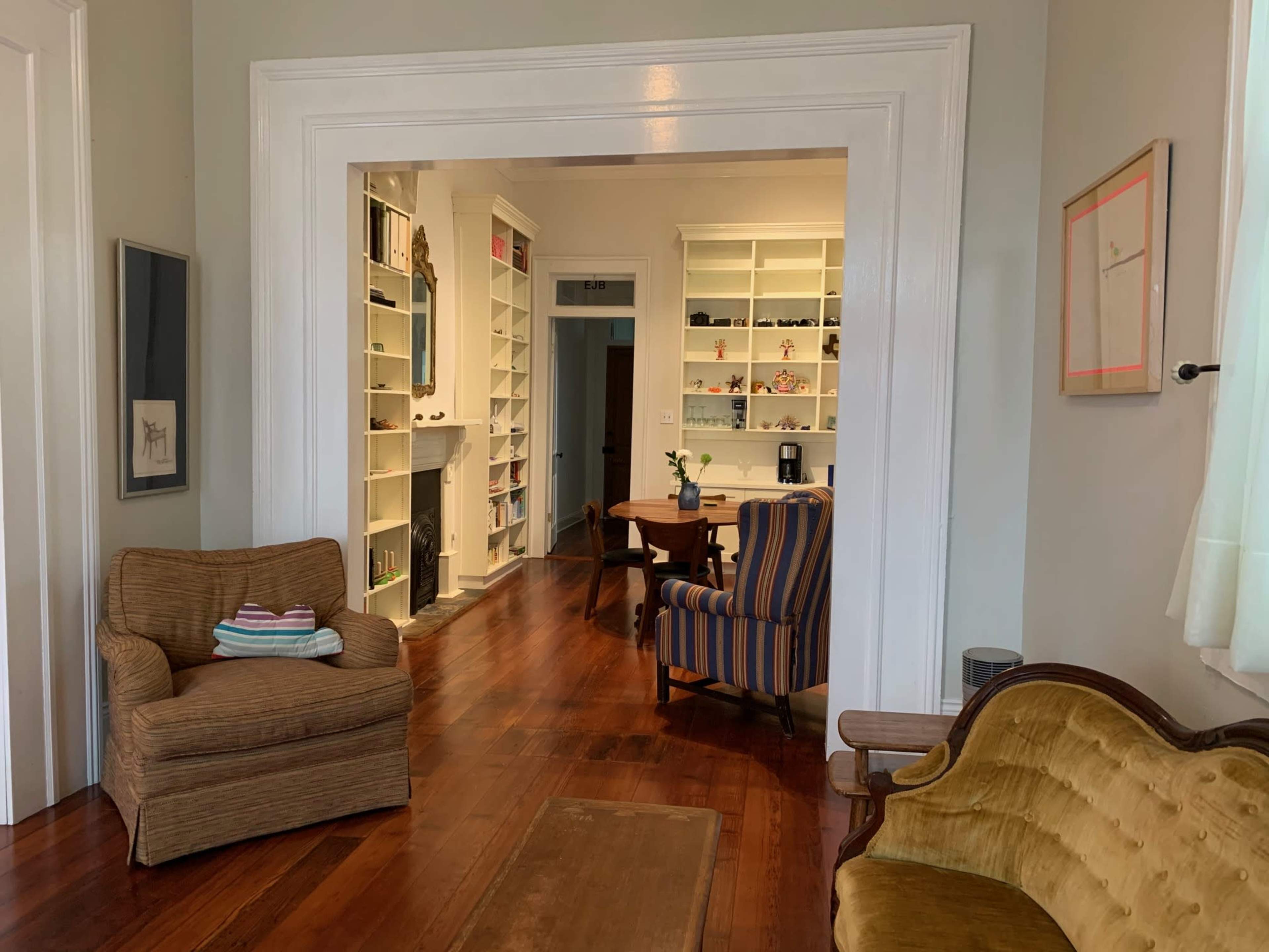The image shows a cozy living room with wooden flooring, featuring a striped armchair, a curved yellow couch, and a small table, leading to a bright, organized study area in the background.