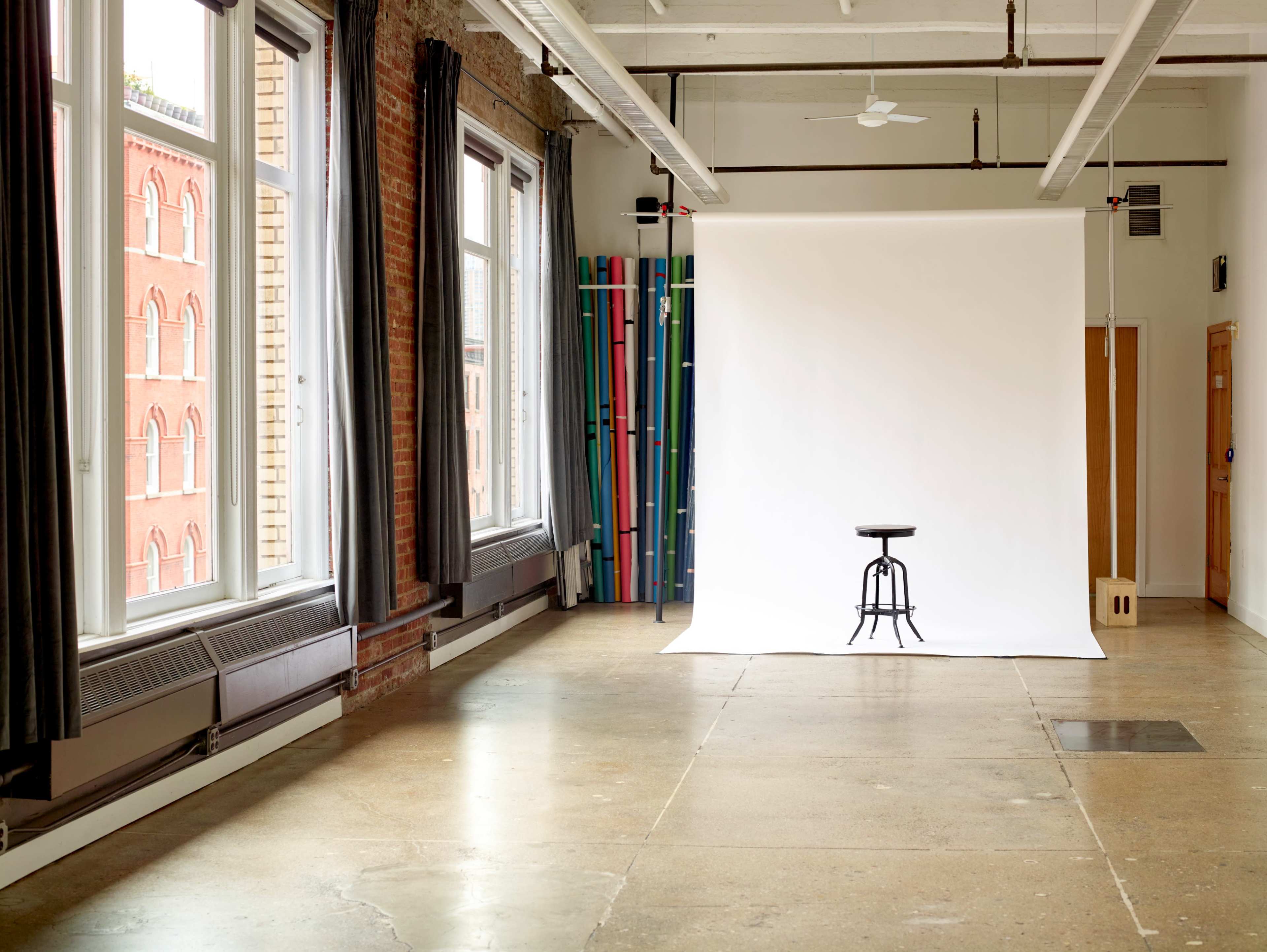 The image shows a spacious room with large windows, featuring a white backdrop, a stool in front of it, and colorful vertical props against the wall.