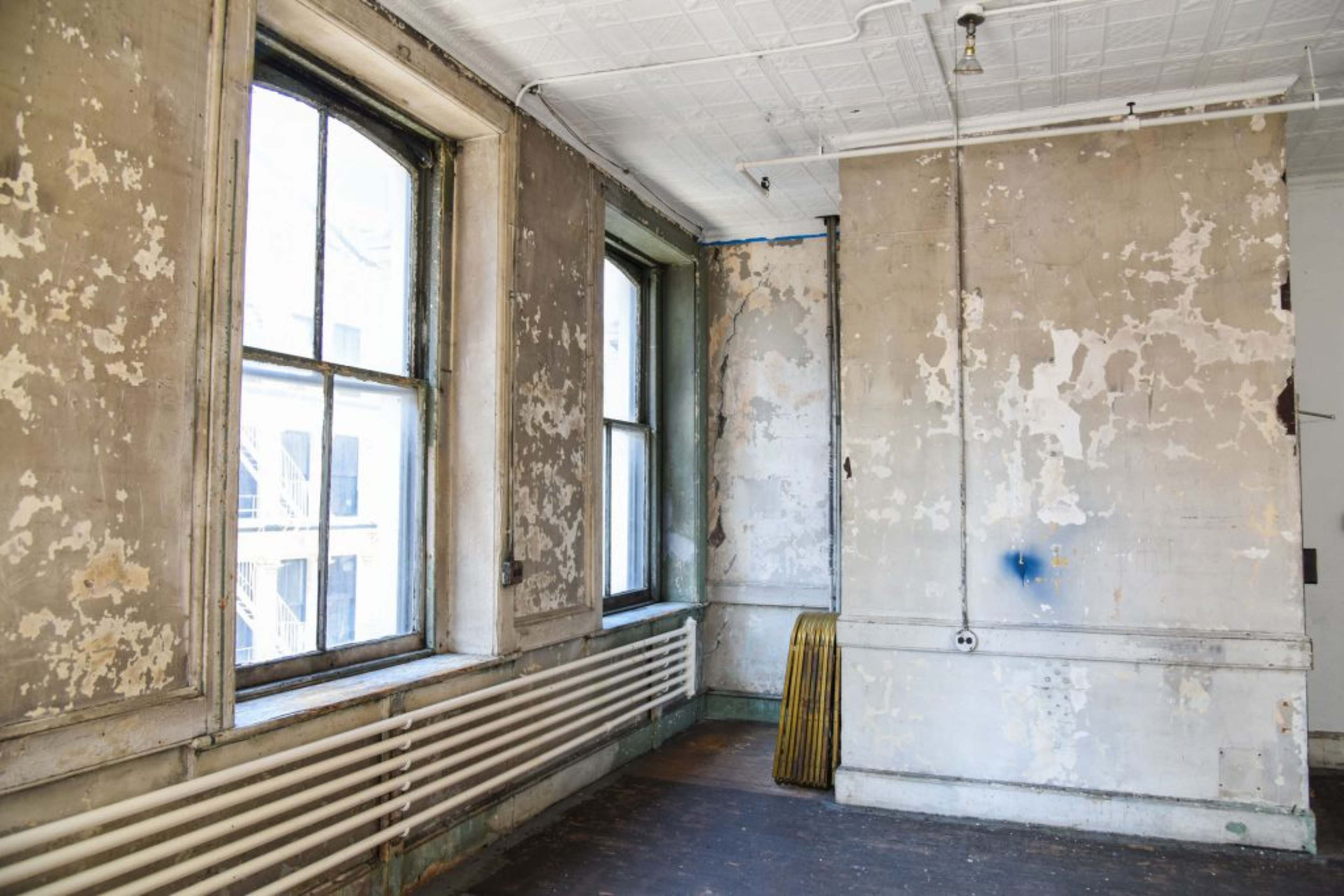 An empty room with peeling paint on the walls, two large windows, and a long radiator along one wall.