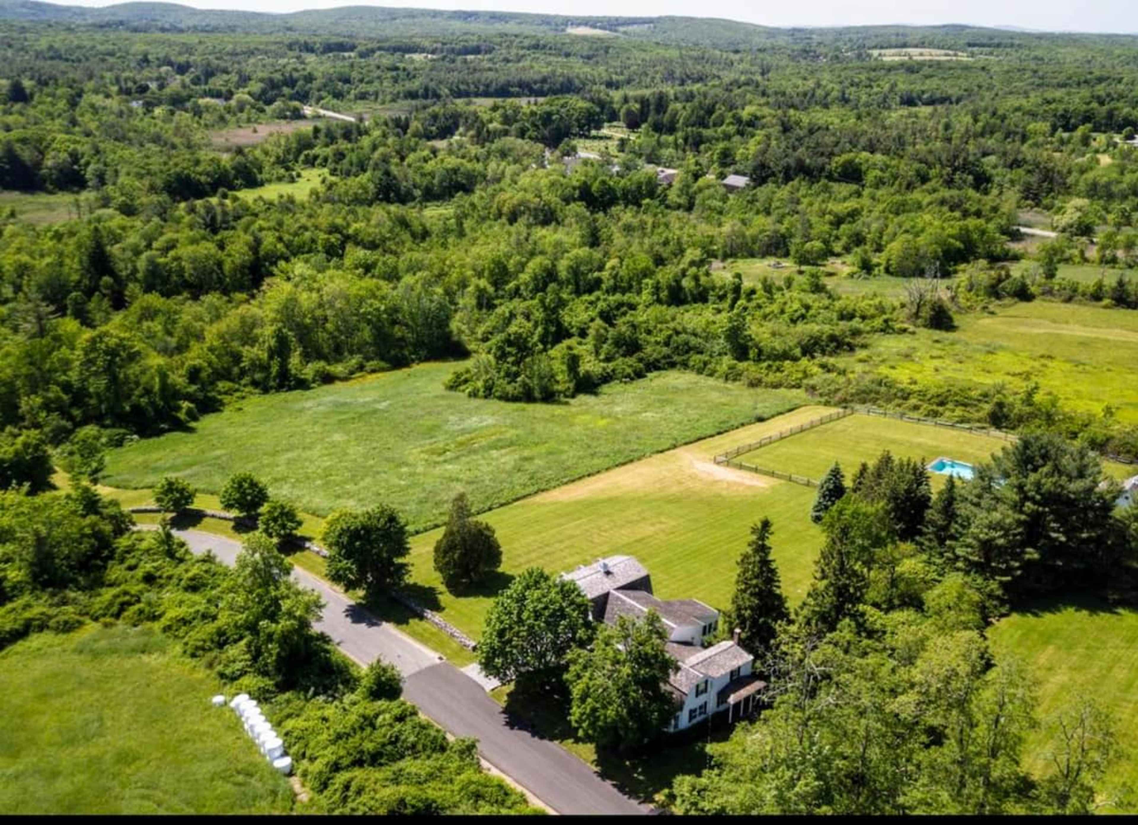 An aerial view shows a spacious, green landscape featuring a large house, well-maintained lawns, and surrounding wooded areas.