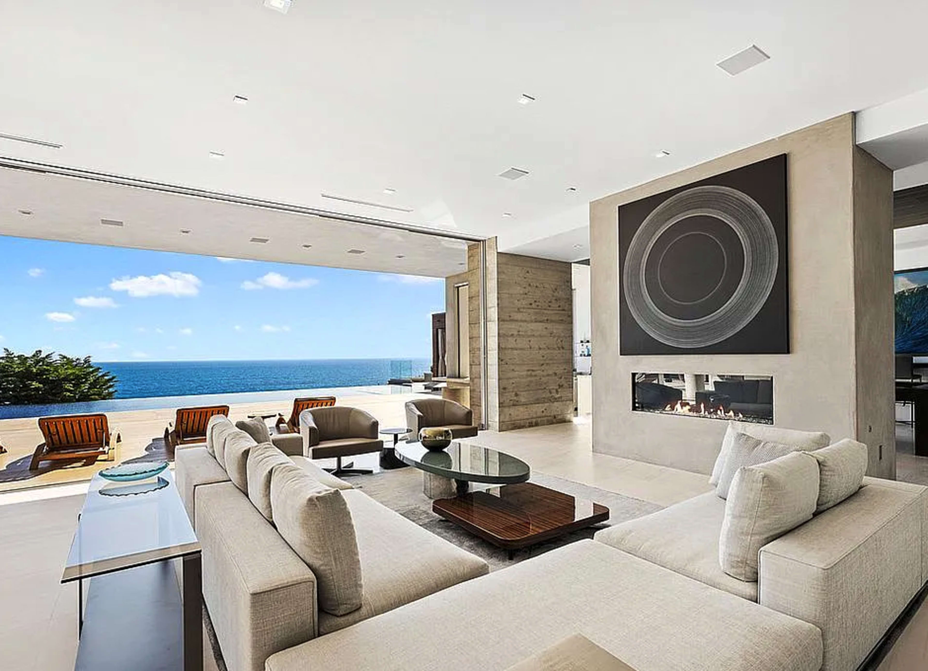 The image shows a modern living room with a large sectional sofa facing a floor-to-ceiling window that opens to a view of the ocean.