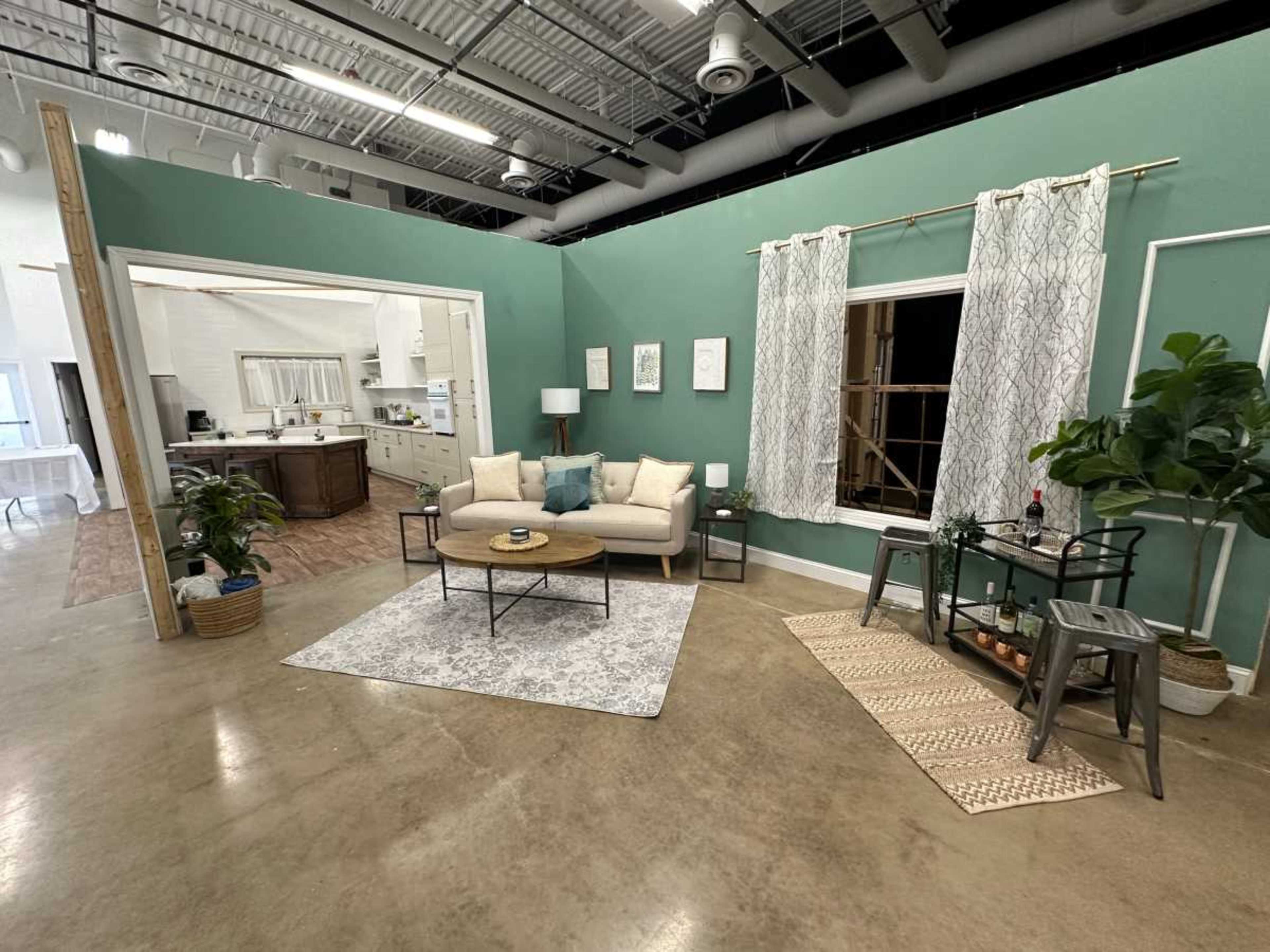 A modern living area with a light-colored sofa, a round coffee table, potted plants, and a decorative rug, bordered by a green wall and a doorway leading to a kitchen.