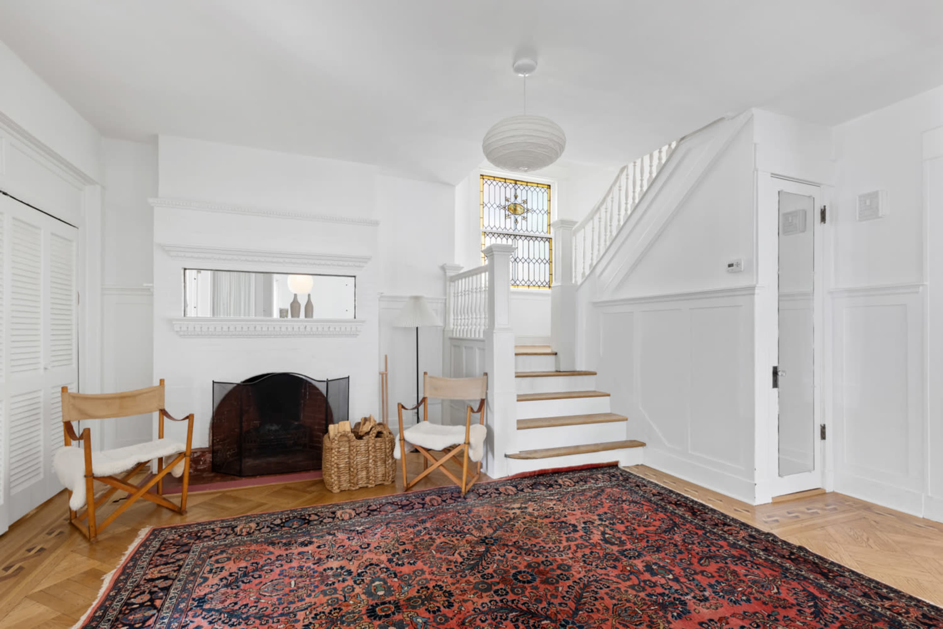 The scene shows a bright, minimalist entryway with a staircase, a fireplace, and a large area rug, complemented by light wooden flooring and white walls.