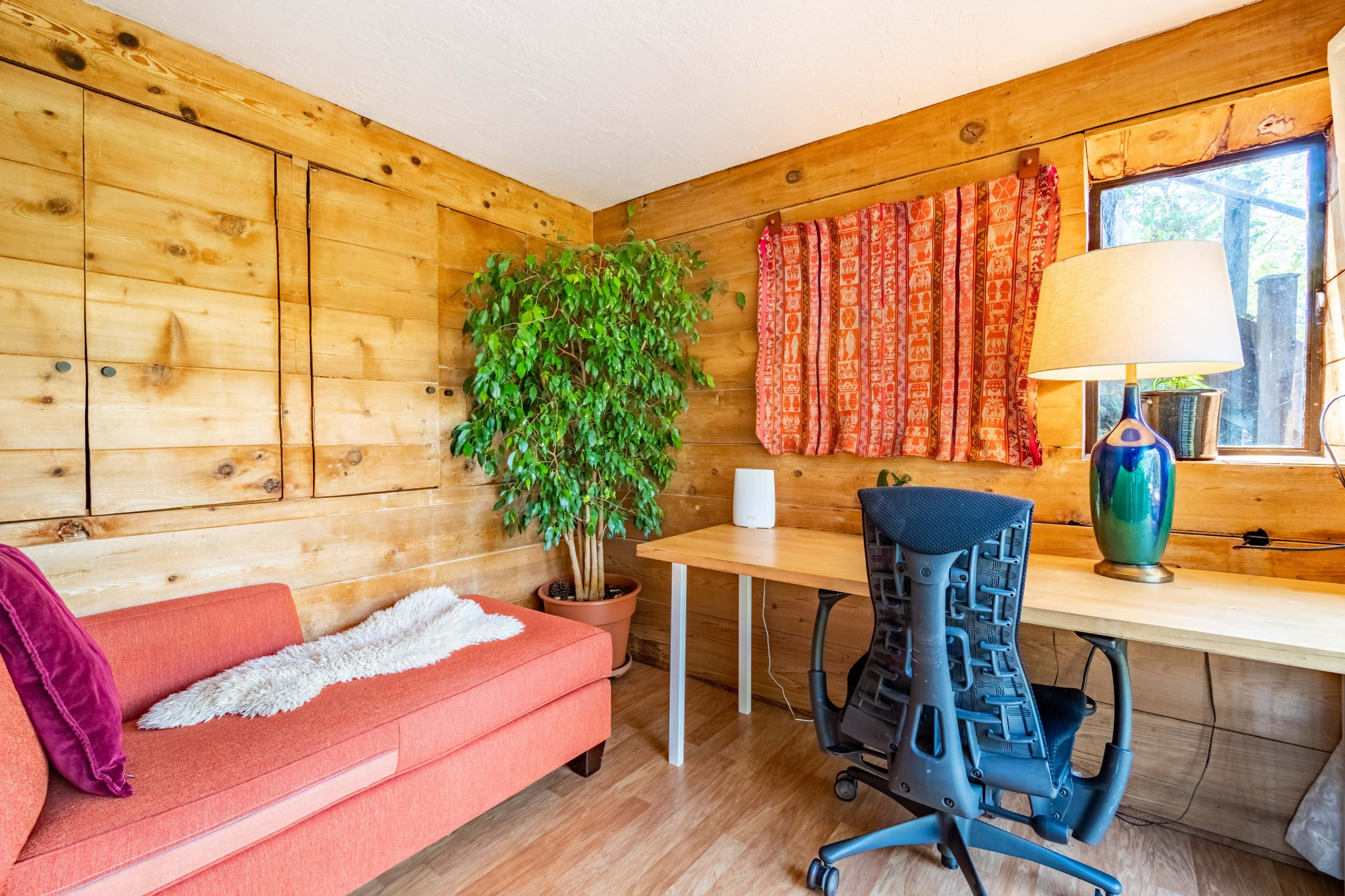A small wooden room featuring a green plant, a desk with a chair, and a red couch with a decorative throw.