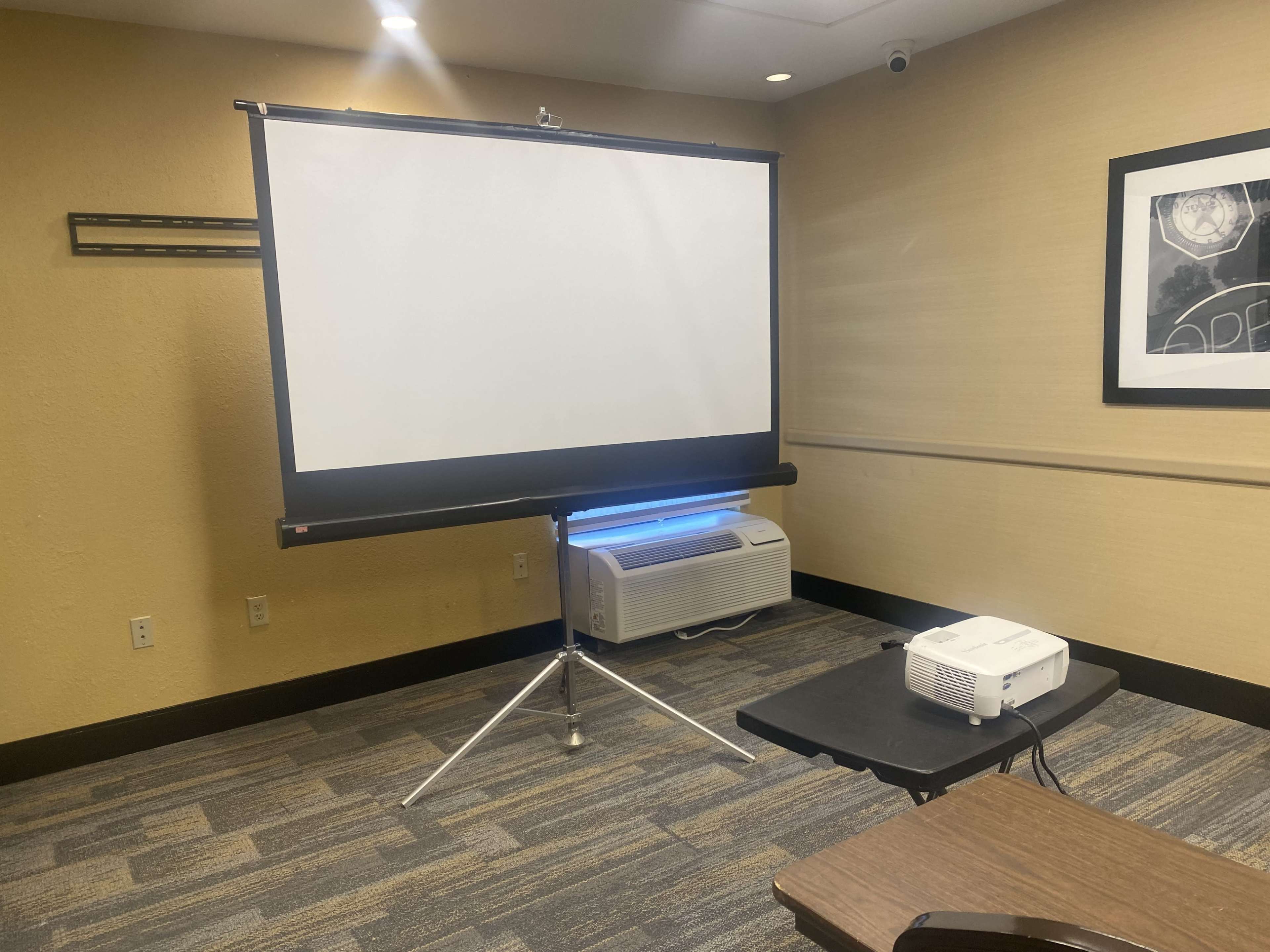 A blank projector screen mounted on a stand next to a small table with a projector in a conference room.