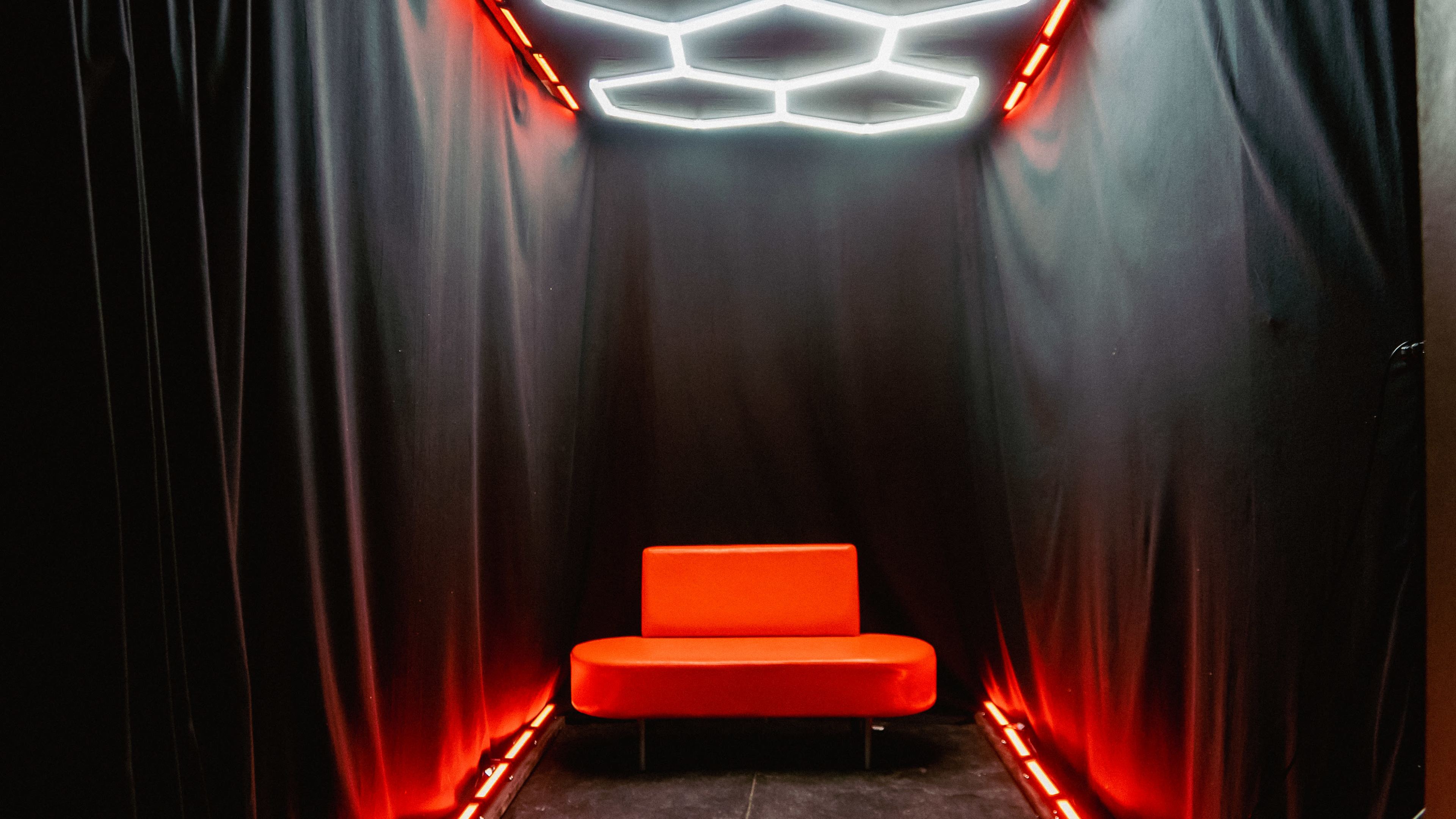 A red couch is positioned in a dimly lit, enclosed space with black curtains and illuminated geometric lighting overhead.