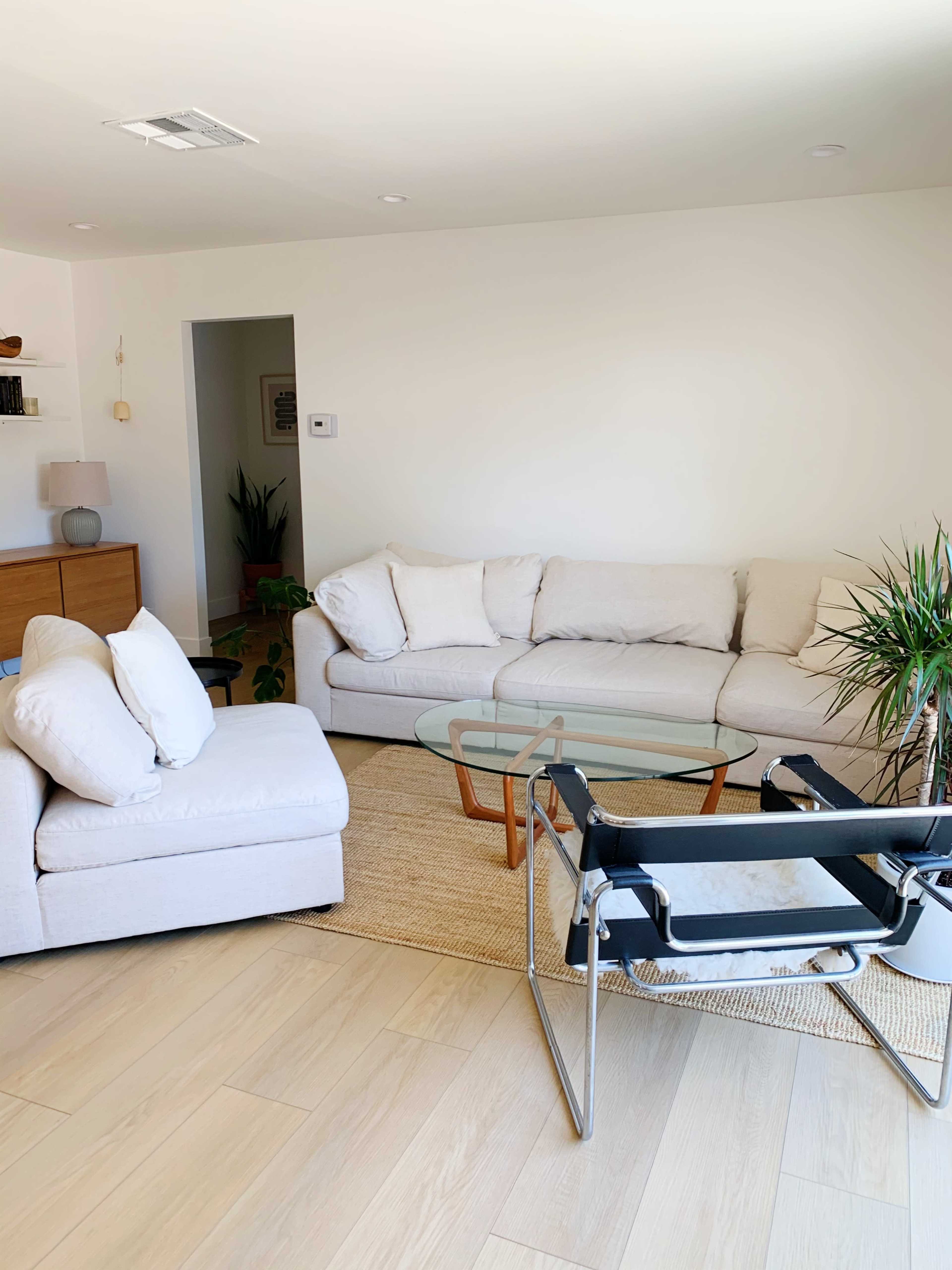 A light-colored living room with a large sectional sofa, a glass coffee table, and a chair, all situated on a beige rug.